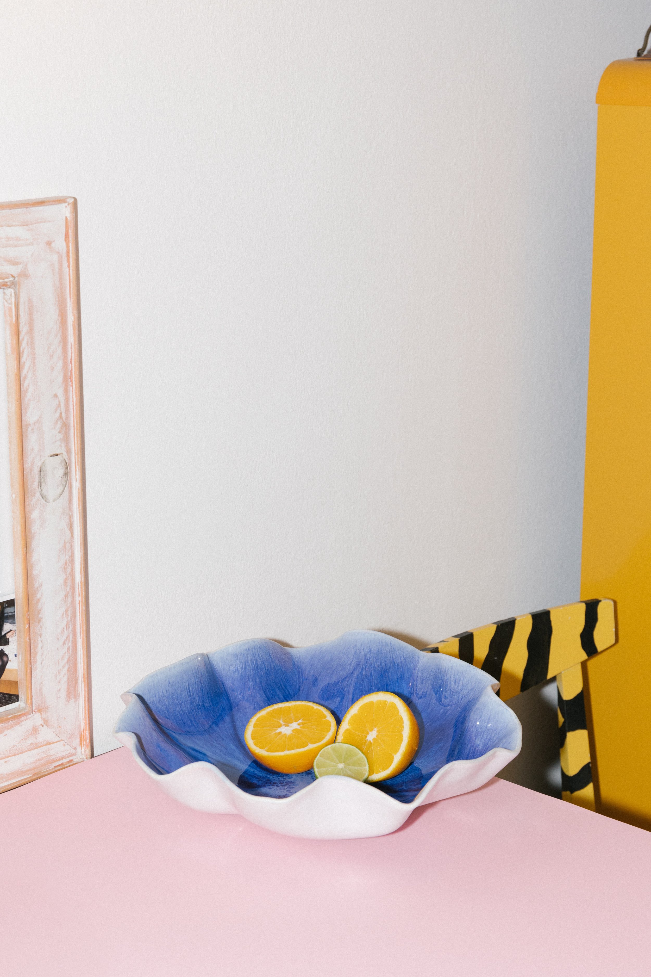 Bunte Glasschale mit halbierten Zitronen und Orangen auf rosa Tisch, Hintergrund mit weißer Wand, gelb-schwarzes gestreiftes Stuhl, gelber Gegenstand.