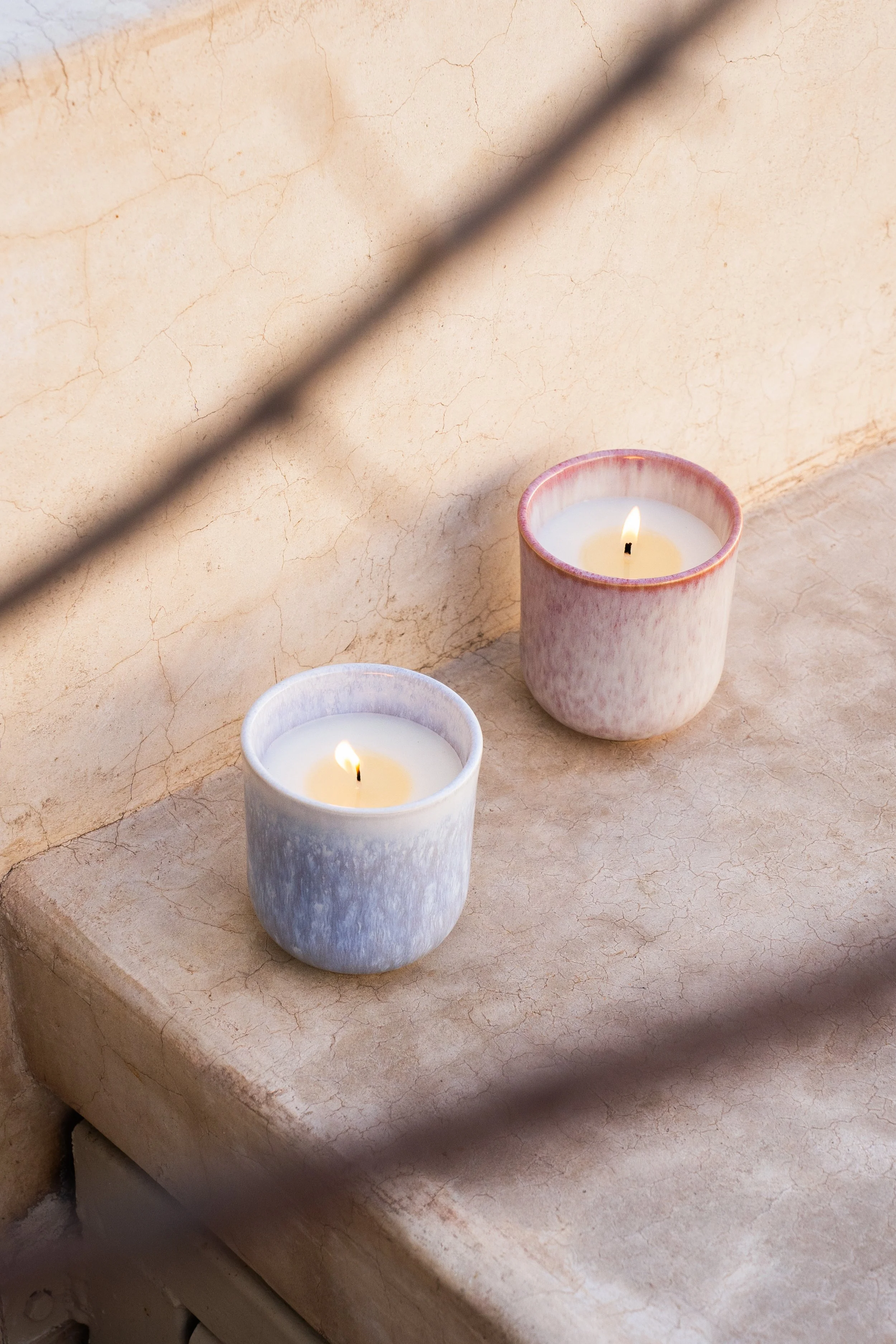 Zwei Kerzen in Kerzenhaltern auf einer Marmoroberfläche, mit einer Mauer im Hintergrund und einer schattigen Linie im Vordergrund.