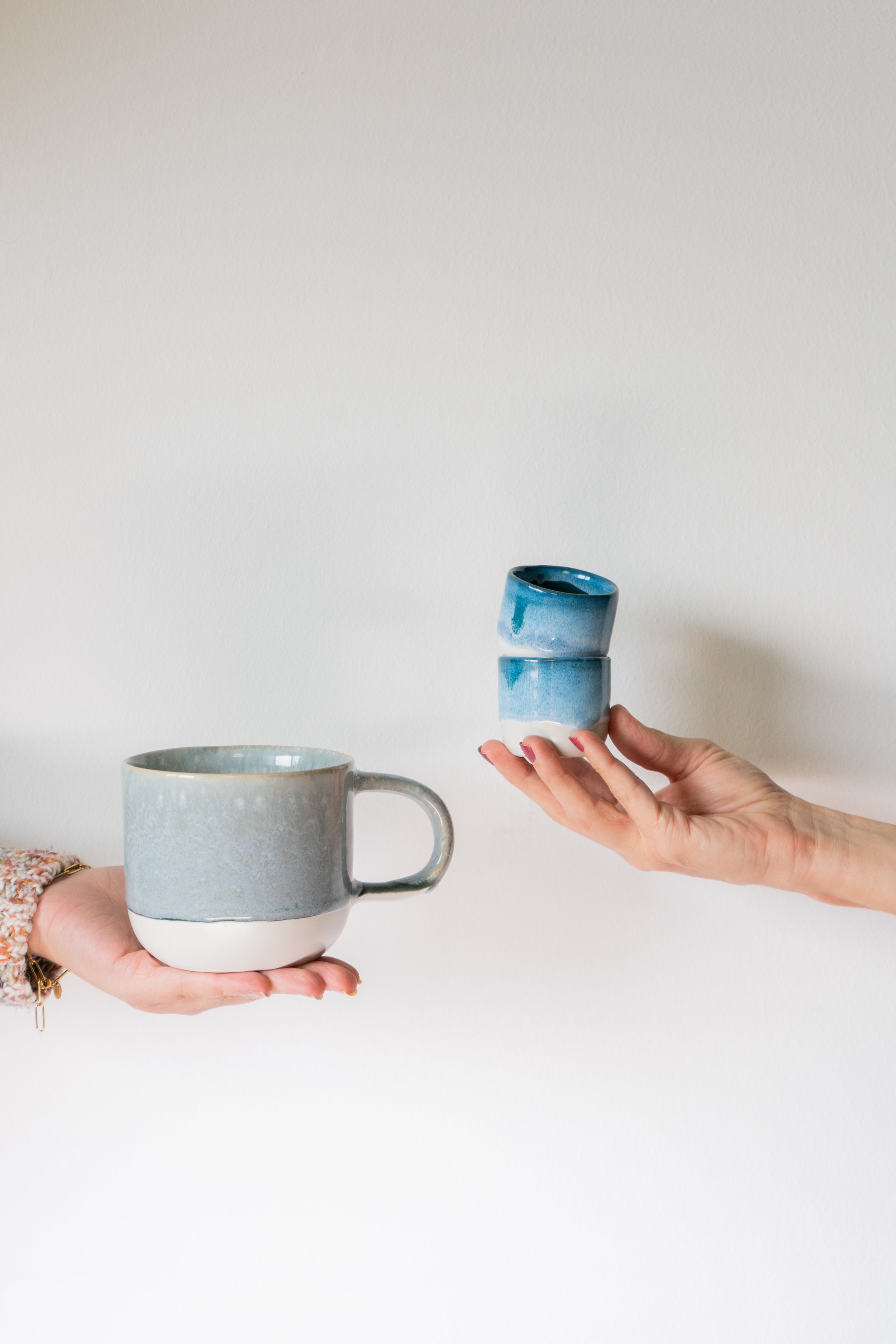 Zwei Hände halten keramische Tassen gegen einen weißen Hintergrund, eine große Tasse auf der linken Seite und zwei kleinere Tassen auf der rechten Seite.