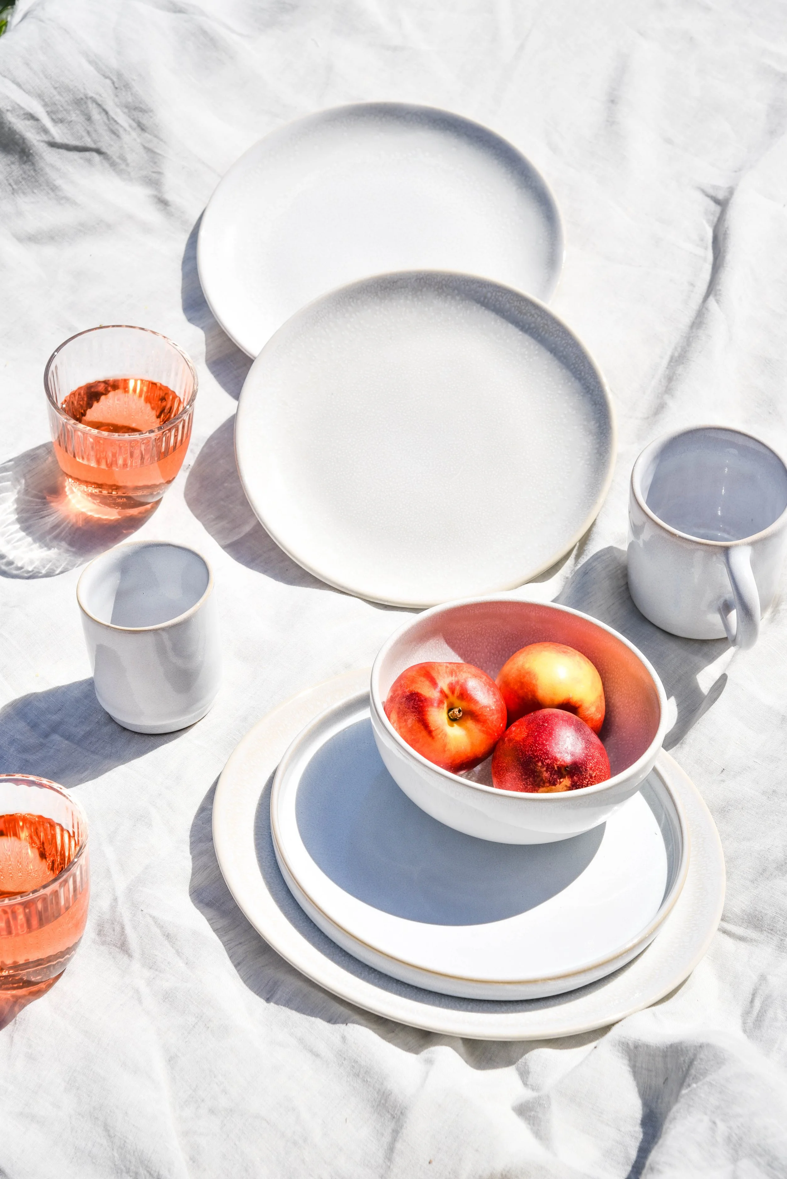 Weiße Speiseteller, Tassen, Gläser und eine Schale mit Äpfeln auf einem weißen Tischtuch im Sonnenlicht.