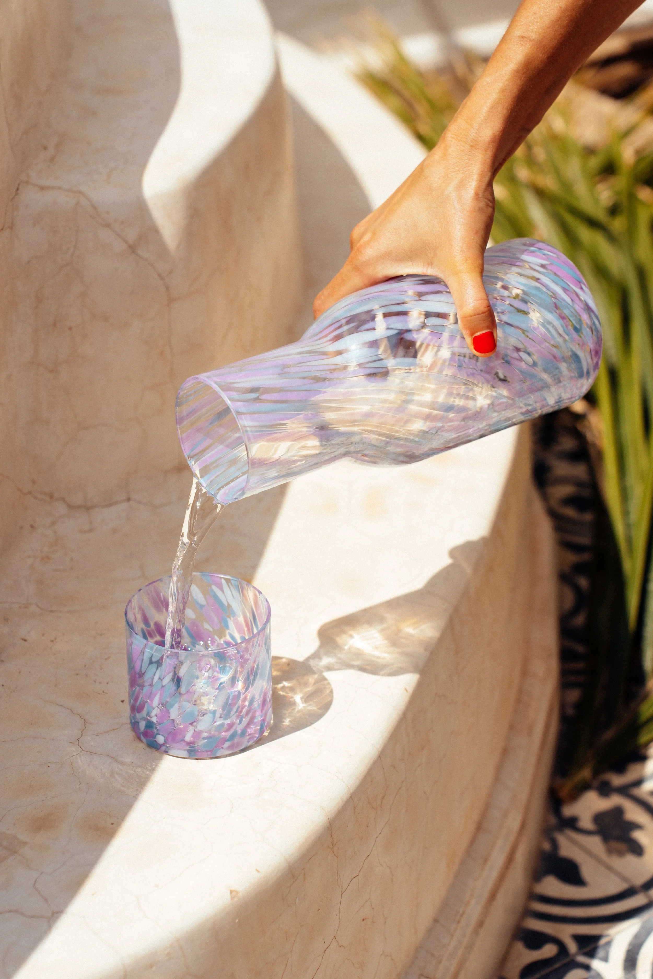 Eine Hand gießt Wasser aus einer bunten, irisierenden Glas Karaffe in ein passendes Glas auf einem hellen, marmorierten Tisch. Im Hintergrund sind grüne Pflanzen sichtbar.