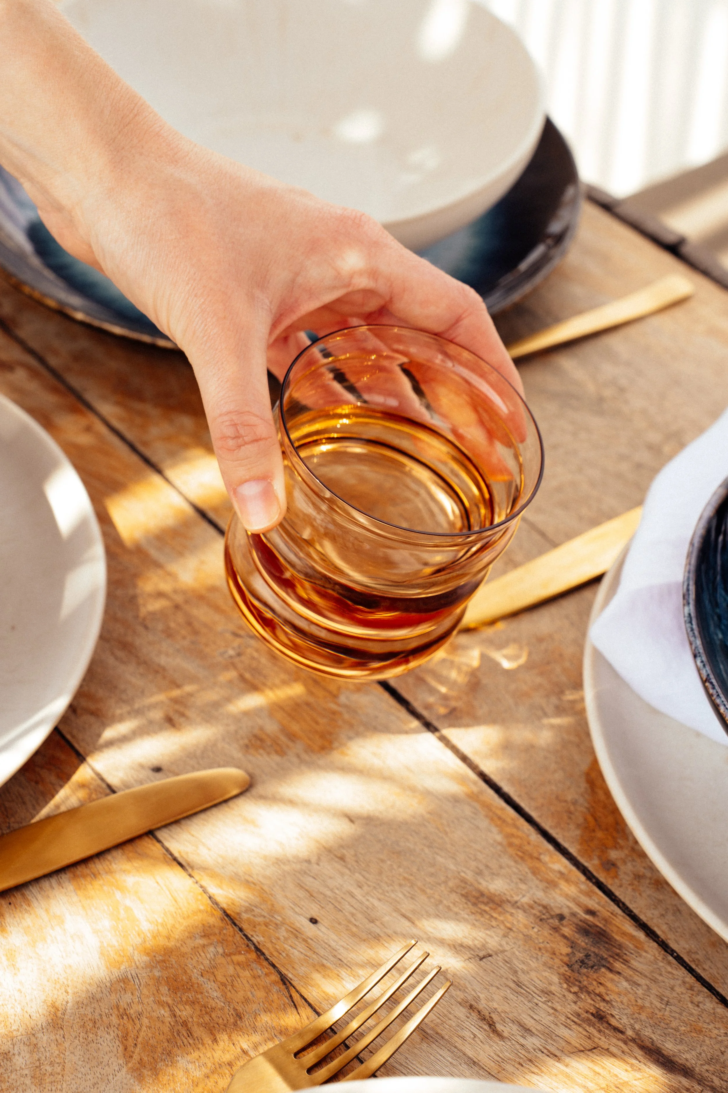 Nahaufnahme einer Hand, die ein Glas mit rosafarbener Flüssigkeit auf einem rustikalen Holztisch hält, umgeben von Goldbesteck und Geschirr.