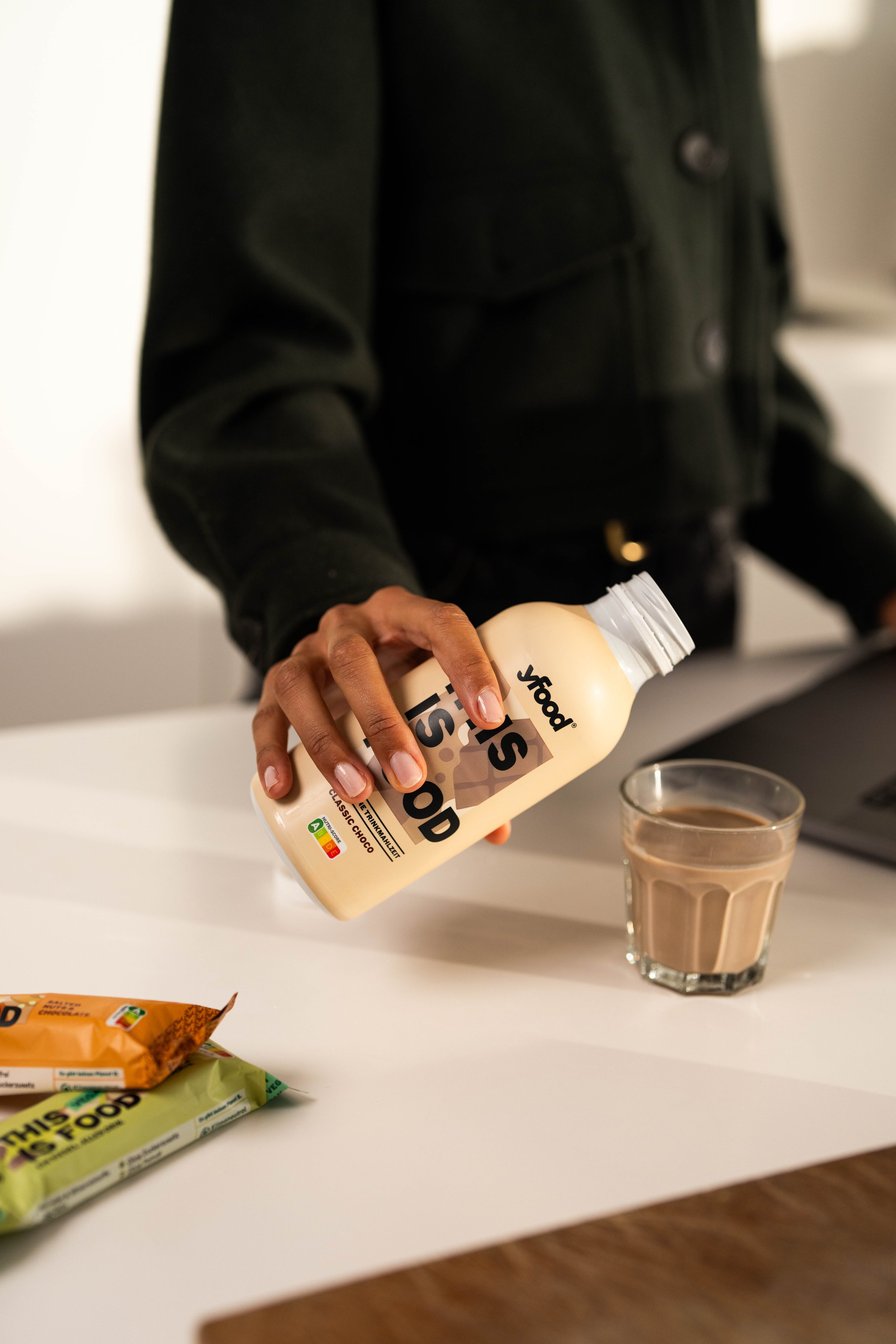 Eine Person in dunkler Jacke hält eine Flasche Essig mit der Aufschrift "Yoop" und ein Glas mit einer braunen Flüssigkeit. Auf dem Tisch liegen zwei Snack-Riegel, auf der rechten Seite ein Laptop.