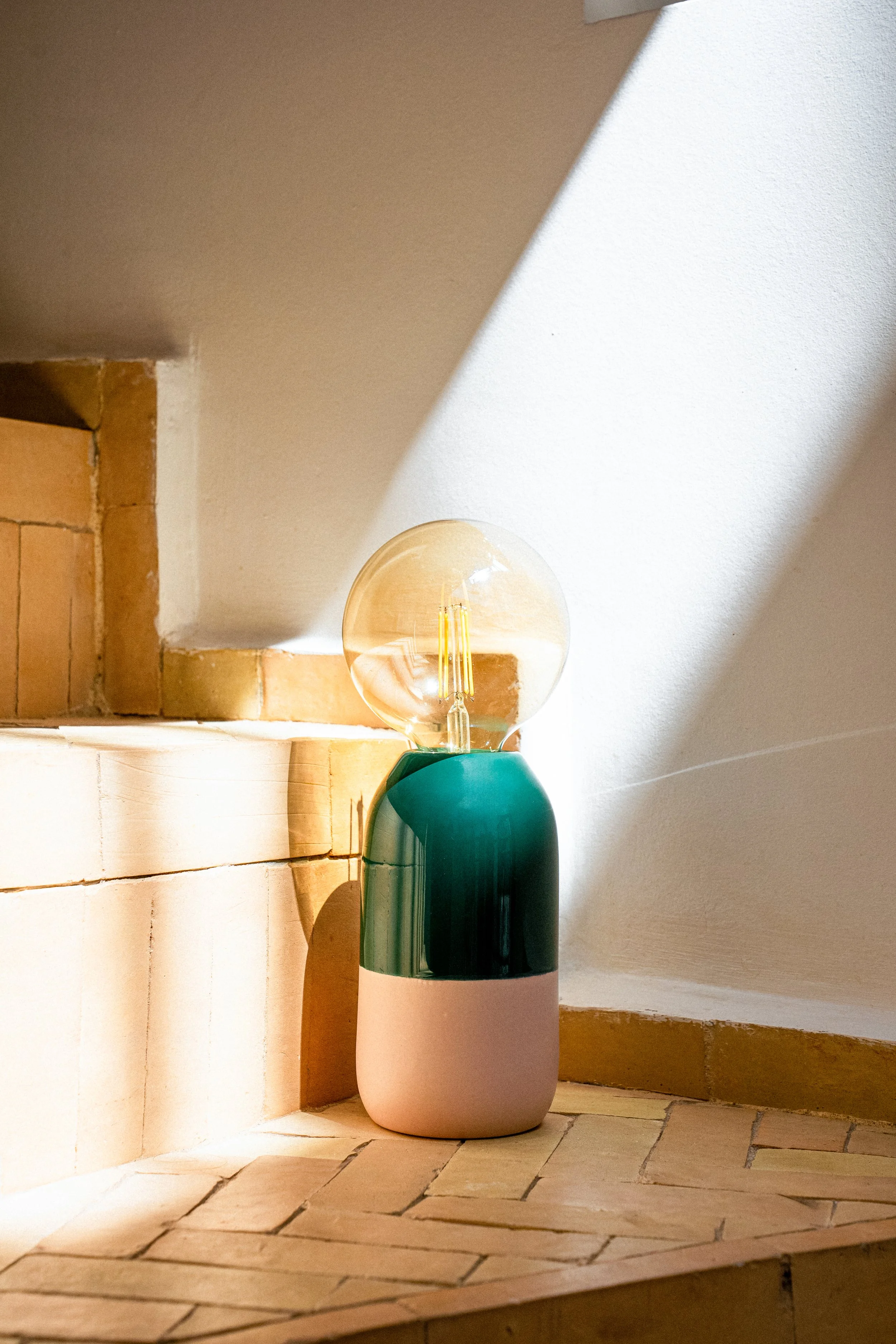 Stehlampe mit einer sichtbaren Glühbirne, auf einem braunen Fliesenboden, im Eck einer weißen Wand.