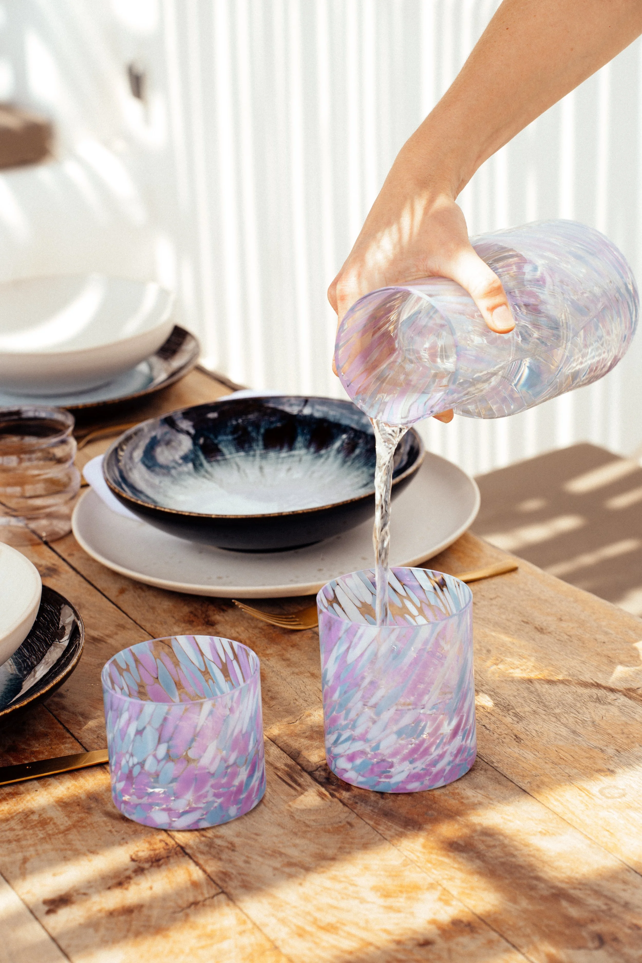 Eine Person gießt Wasser aus einer irisierten Glas Karaffe in ein ähnliches Glas auf einem Holztisch, umgeben von Geschirr und Besteck.