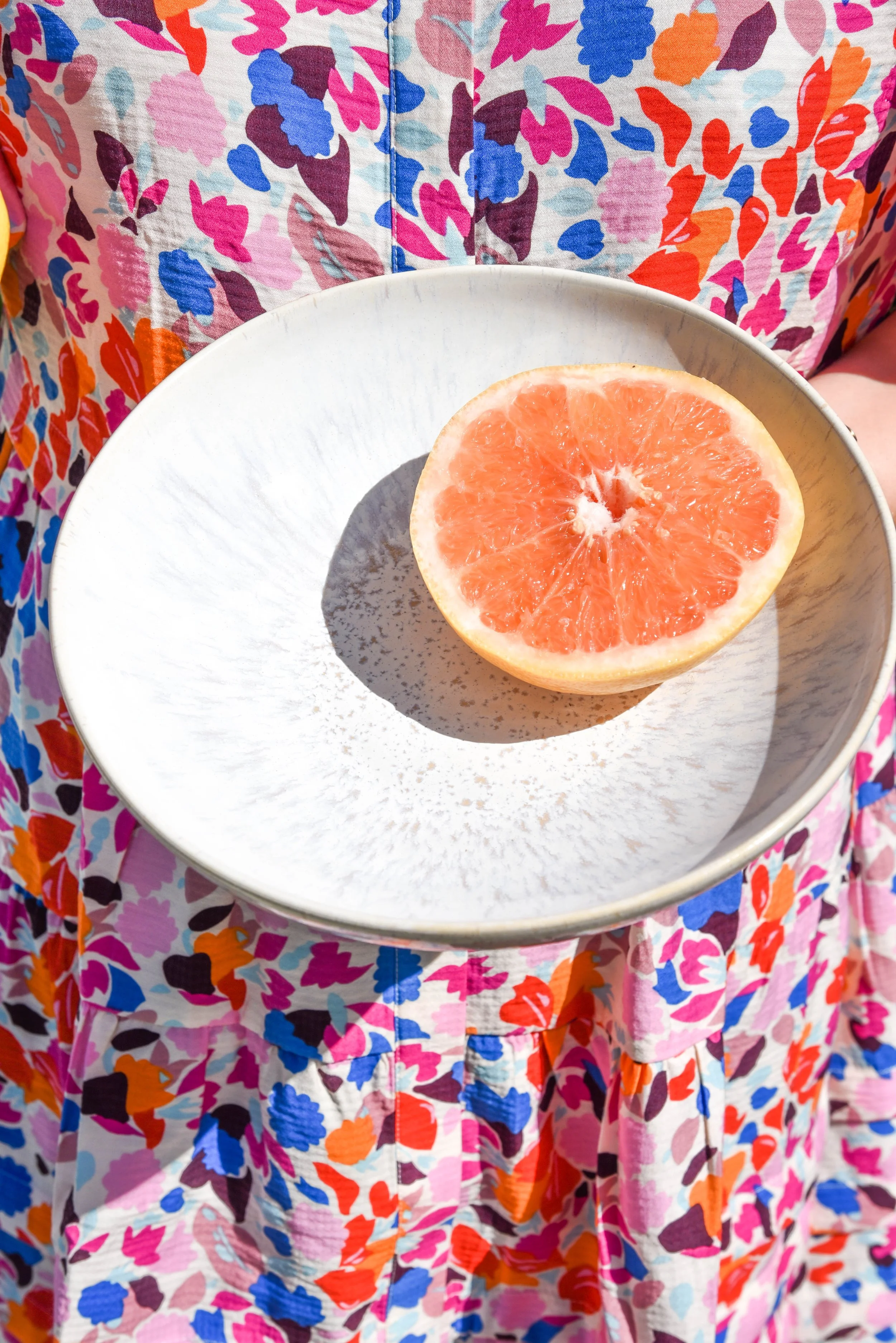 Eine Person in einem bunten, blumenmusterhaften Kleid hält einen weißen Teller mit einer halbierten Grapefruit