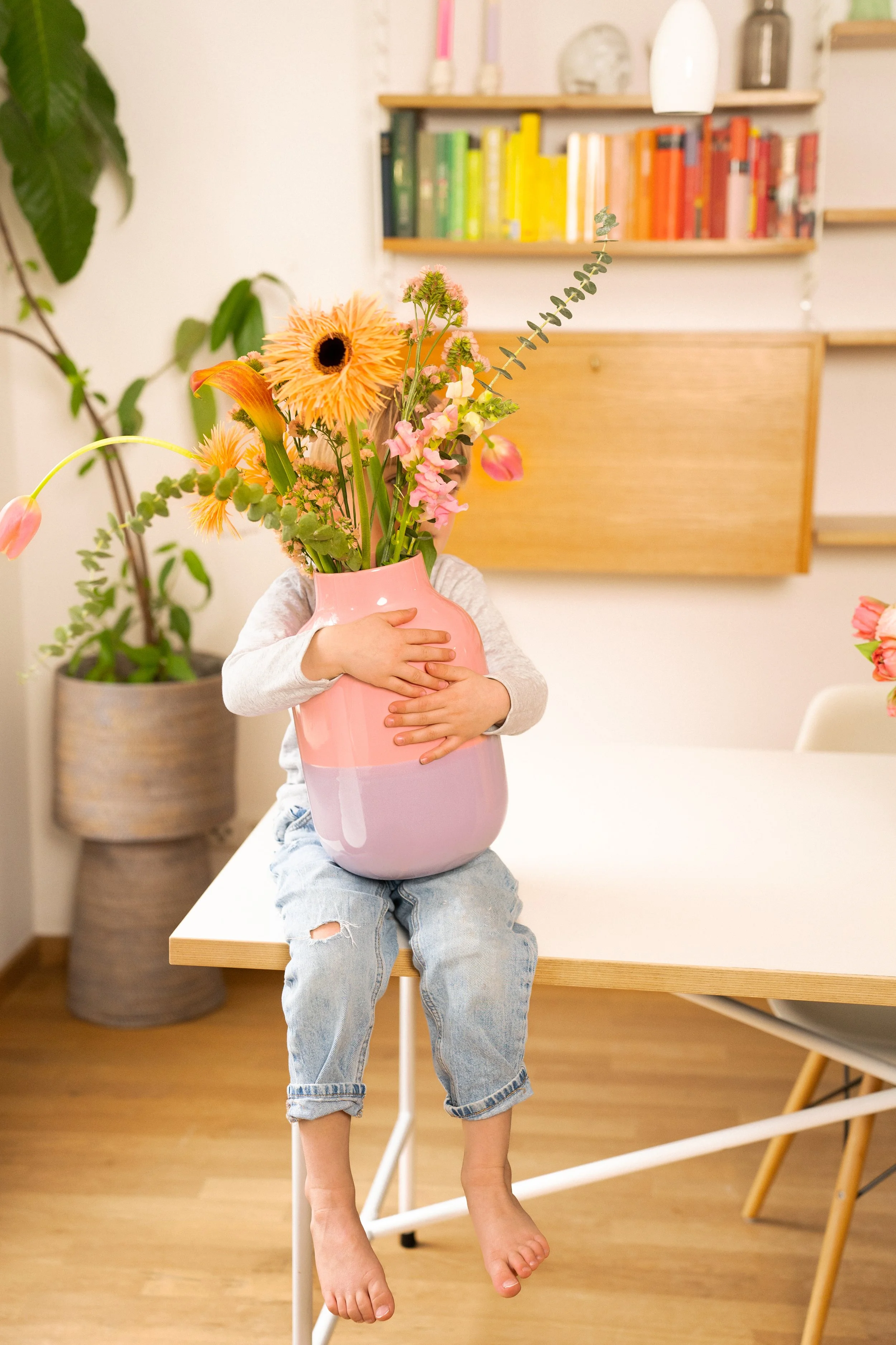 Ein Kind sitzt auf einem Tisch mit einem großen pinken Blumenvasen, das mit bunten Blumen gefüllt ist. Das Kind hat die Arme um die Vase gelegt, die das Gesicht verbirgt. Im Hintergrund sind bunte Bücherregale und Pflanzen im Raum.