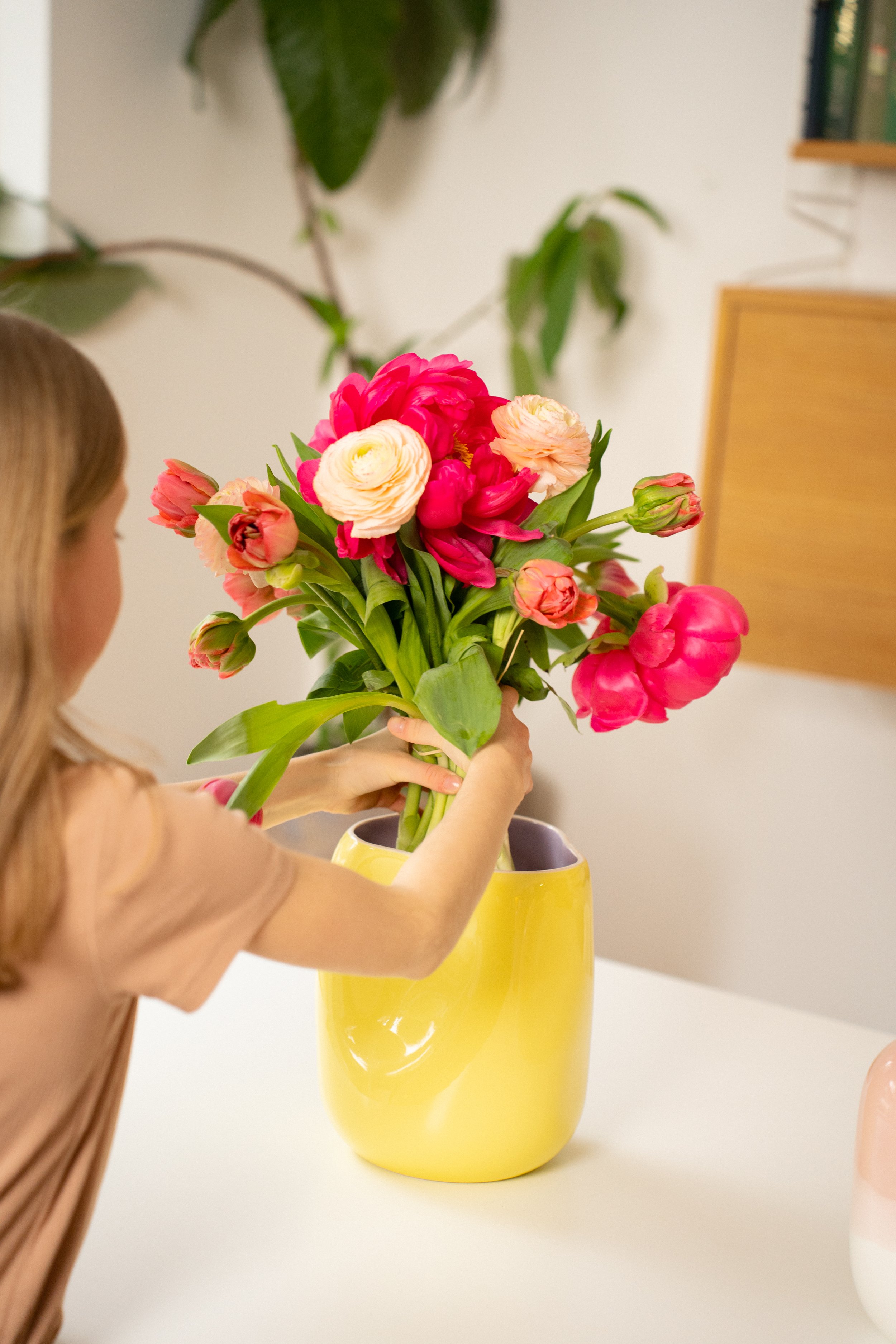 Ein Mädchen, das einen Blumenstrauß mit bunten Tulpen in einem gelben Vasen auf einem weißen Tisch hält.
