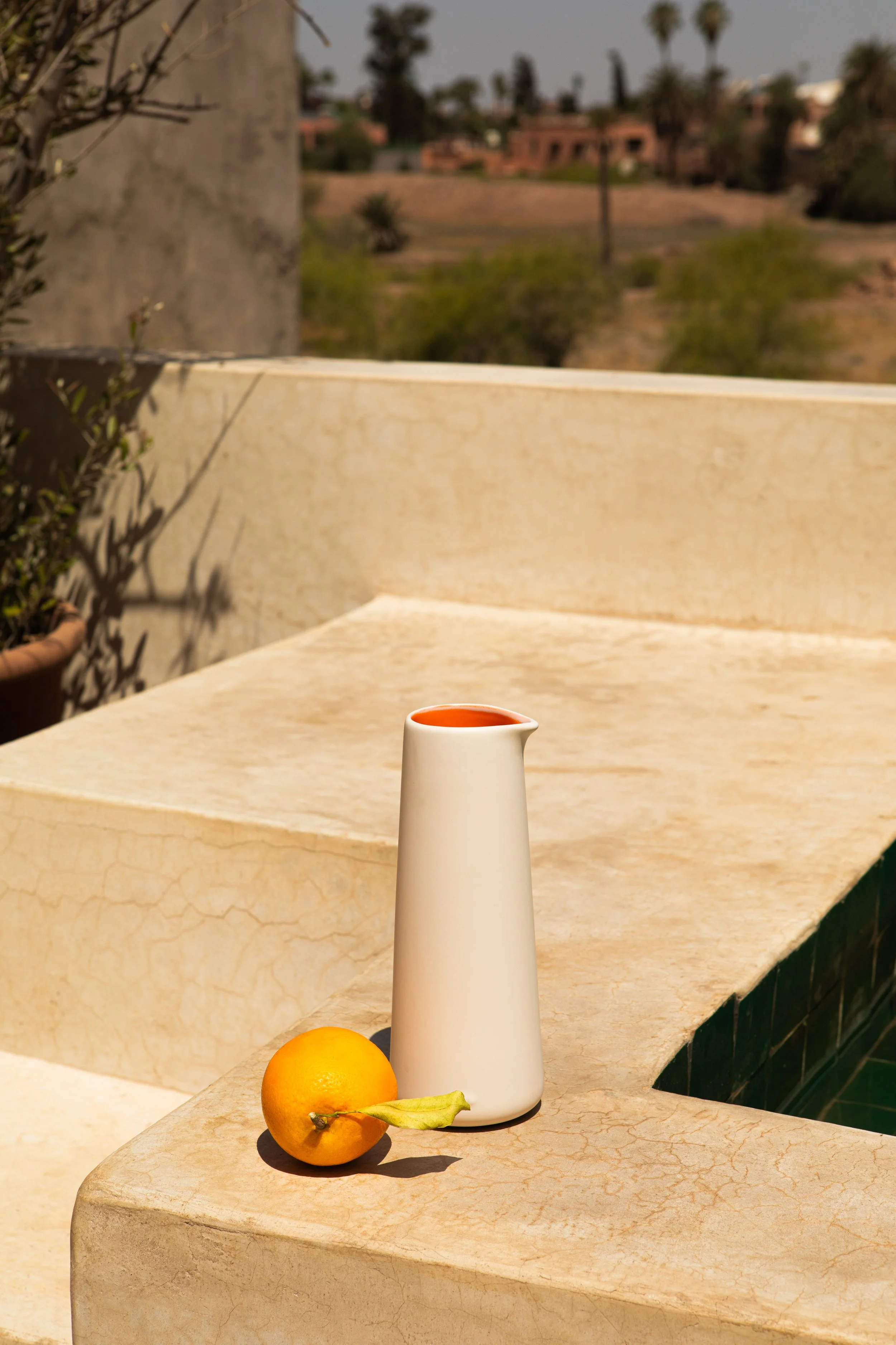 Eine weiße Keramikvase und eine Orange liegen auf einer steinernen Terrasse im Sonnenlicht, im Hintergrund eine Wüstenlandschaft mit Palmen und Gebäuden.