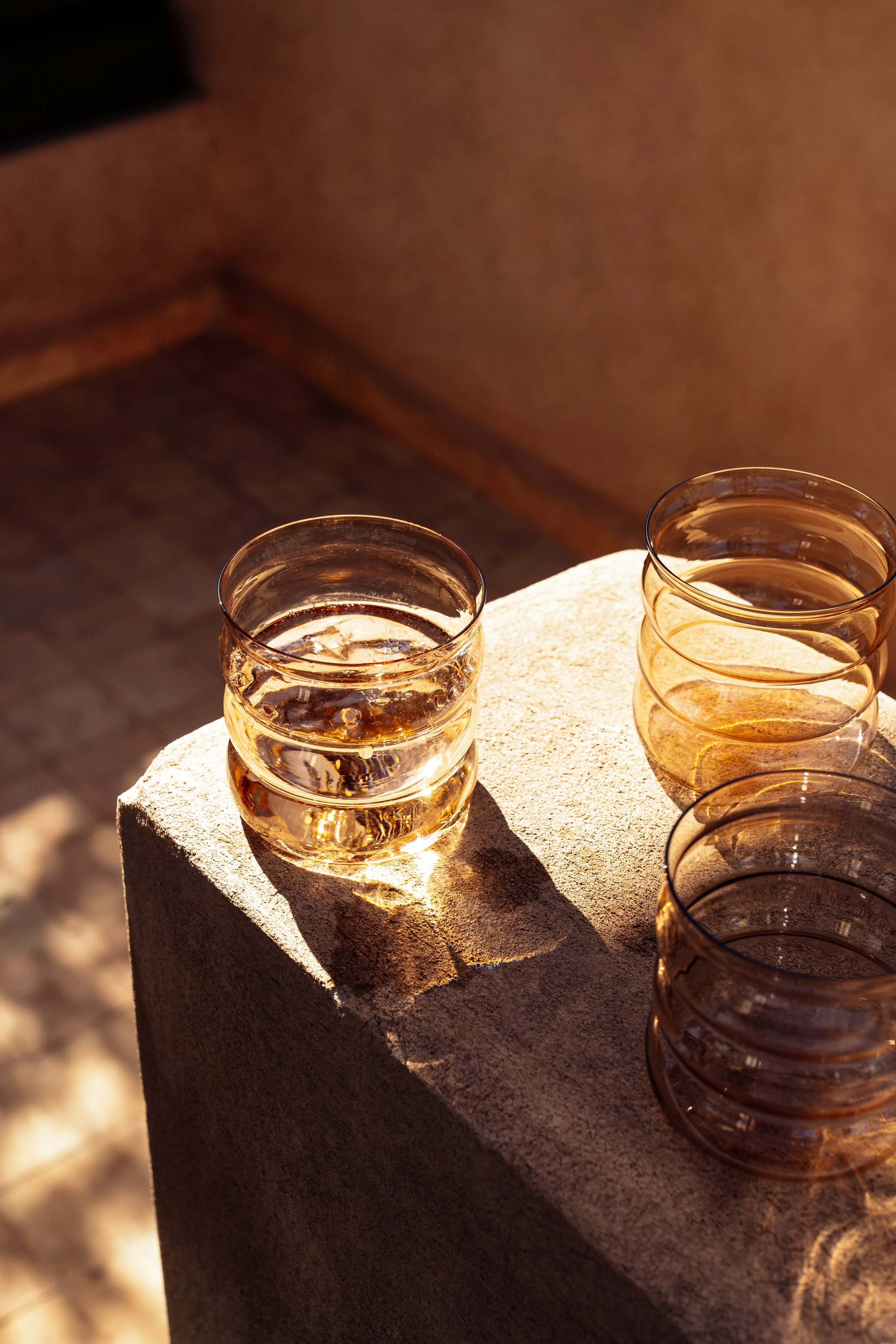 Vier Gläser mit einem goldbraunen Getränk auf einem Steinblock im Sonnenlicht, Schatten auf dem Stein.