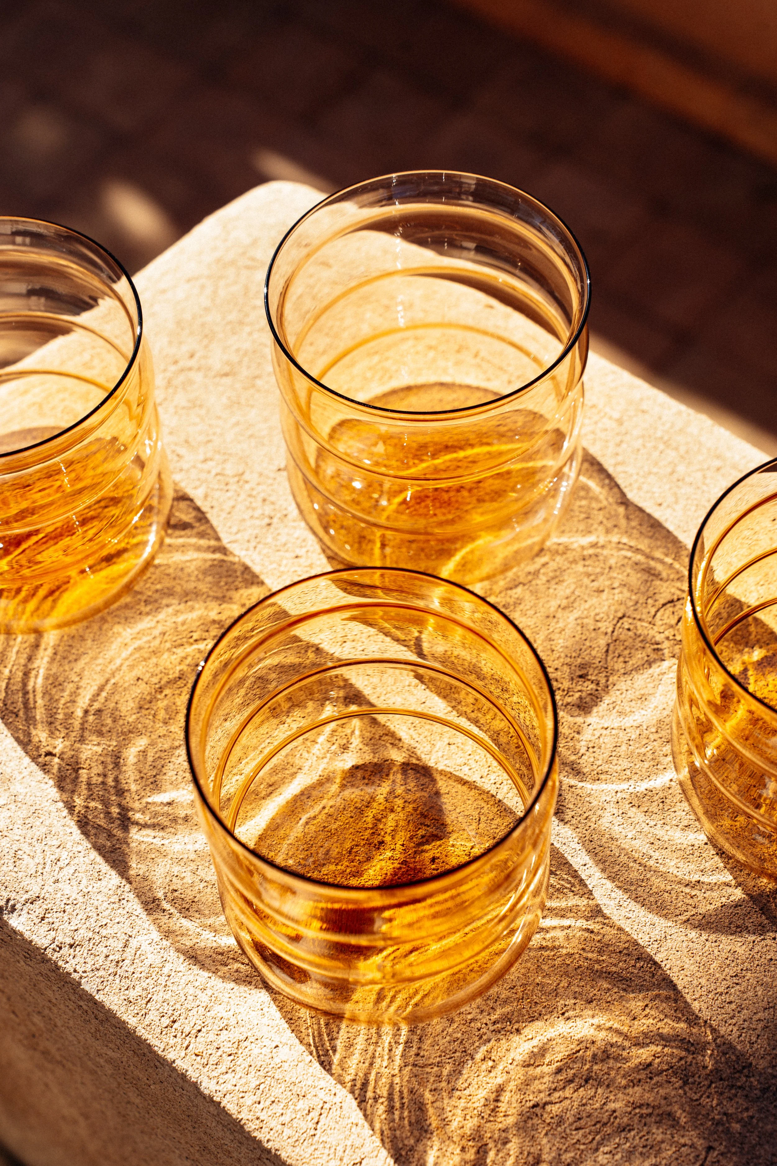 Fünf orangefarbene Gläser mit einem Getränk, auf einem hellen Steinblock, Sonnenlicht wirft Schatten.