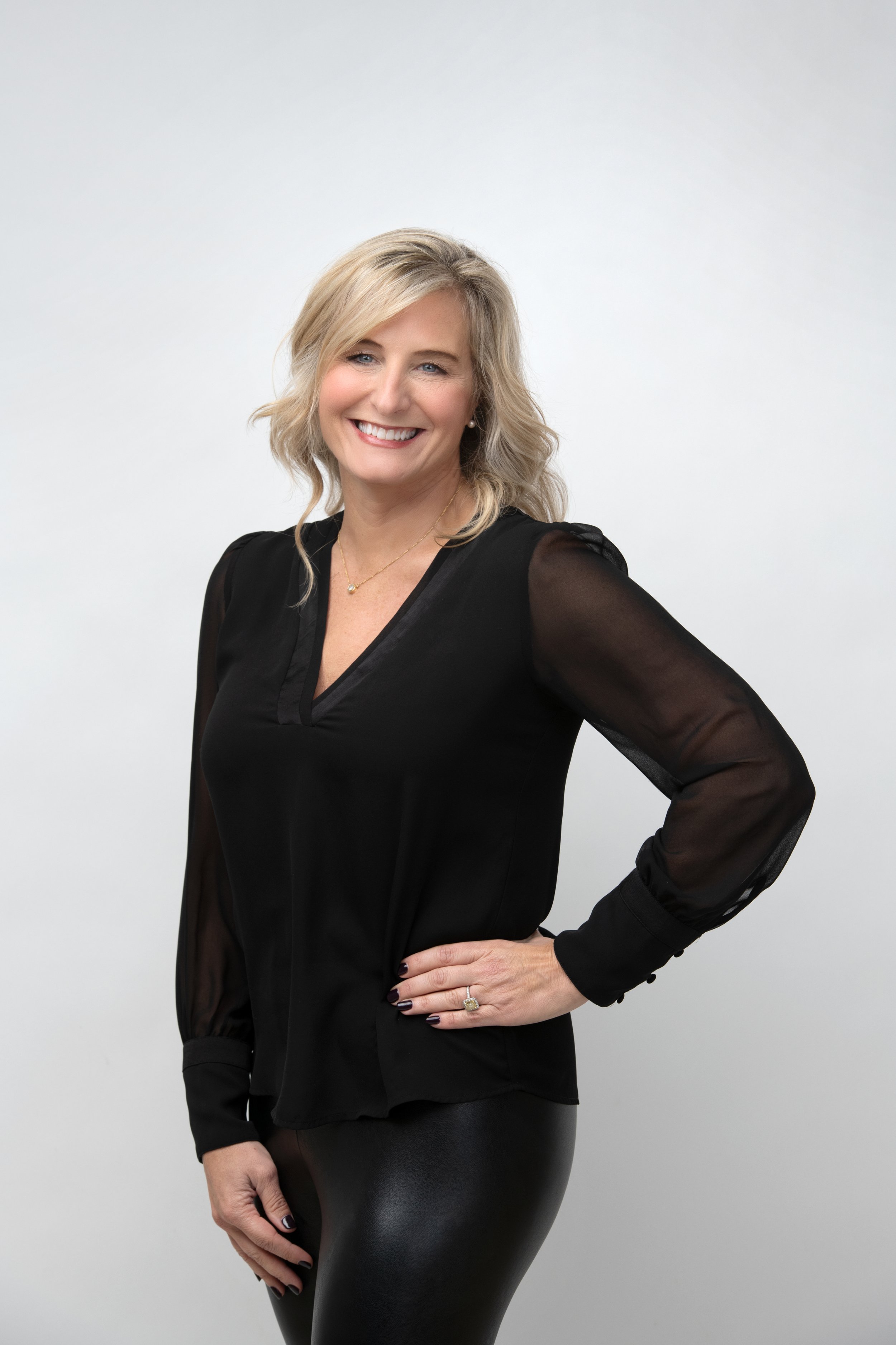 A smiling woman with blonde hair, wearing a black blouse with sheer sleeves and black pants, standing against a plain white background.