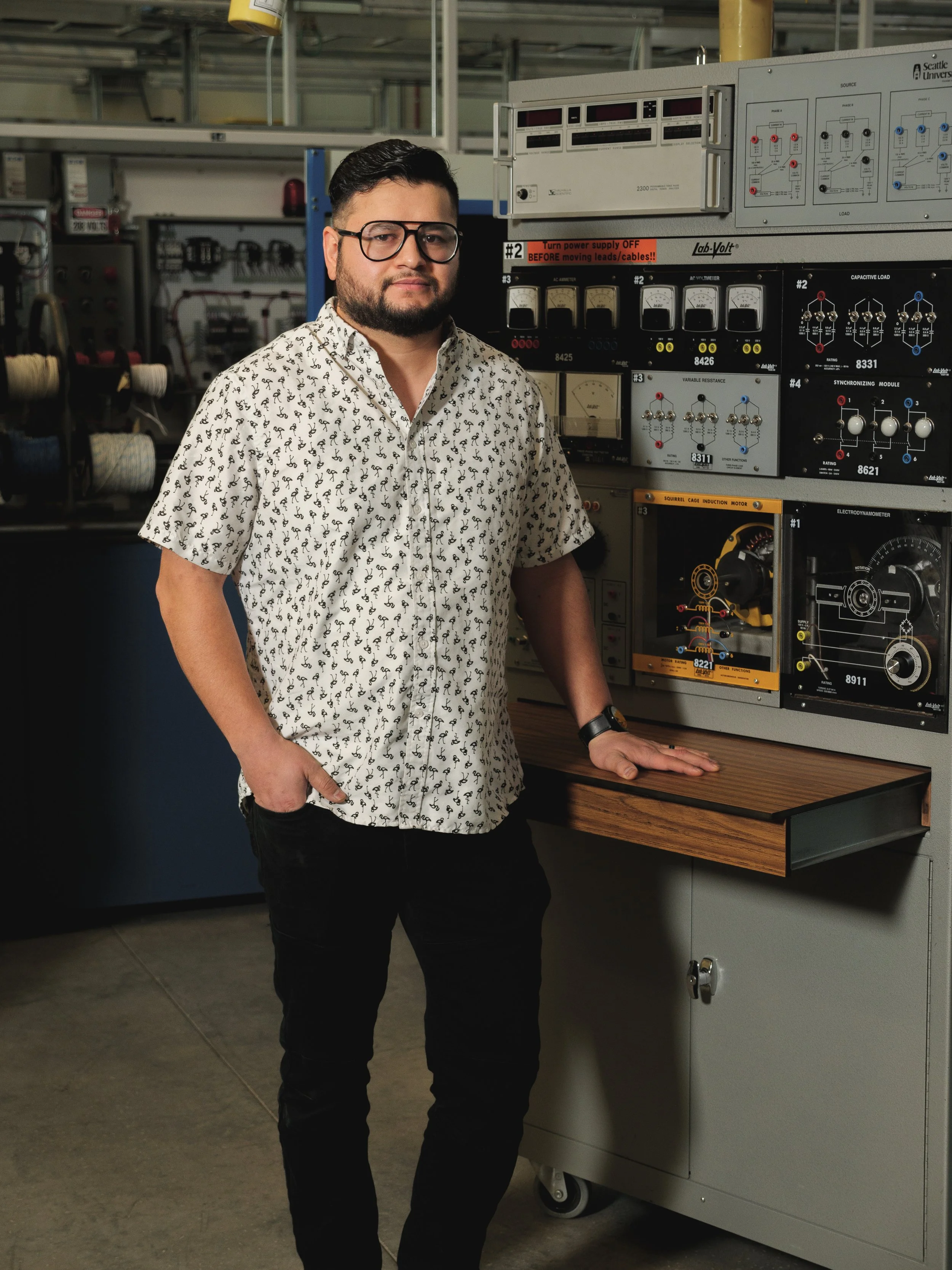 A man with glasses and a beard, wearing a patterned short-sleeve shirt, stands beside a laboratory workbench with electronic equipment, in a technical lab or workshop.