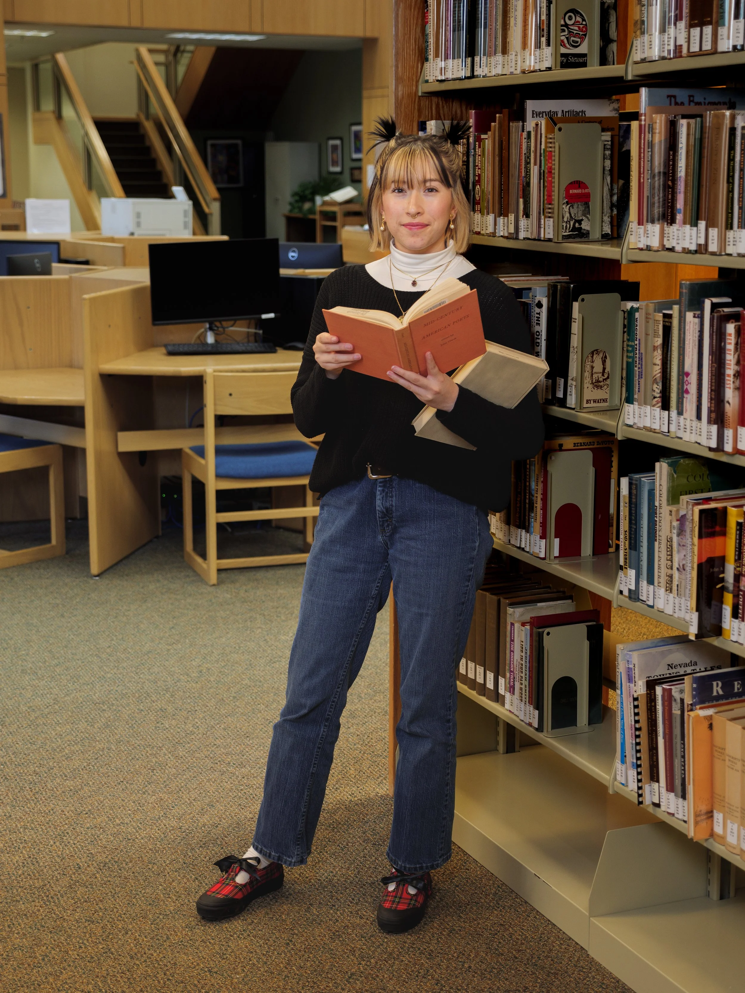 A young woman with light skin and blonde hair styled in pigtails wearing a black sweater, white turtleneck, blue jeans, and plaid shoes, holding an open book while standing in a library aisle.