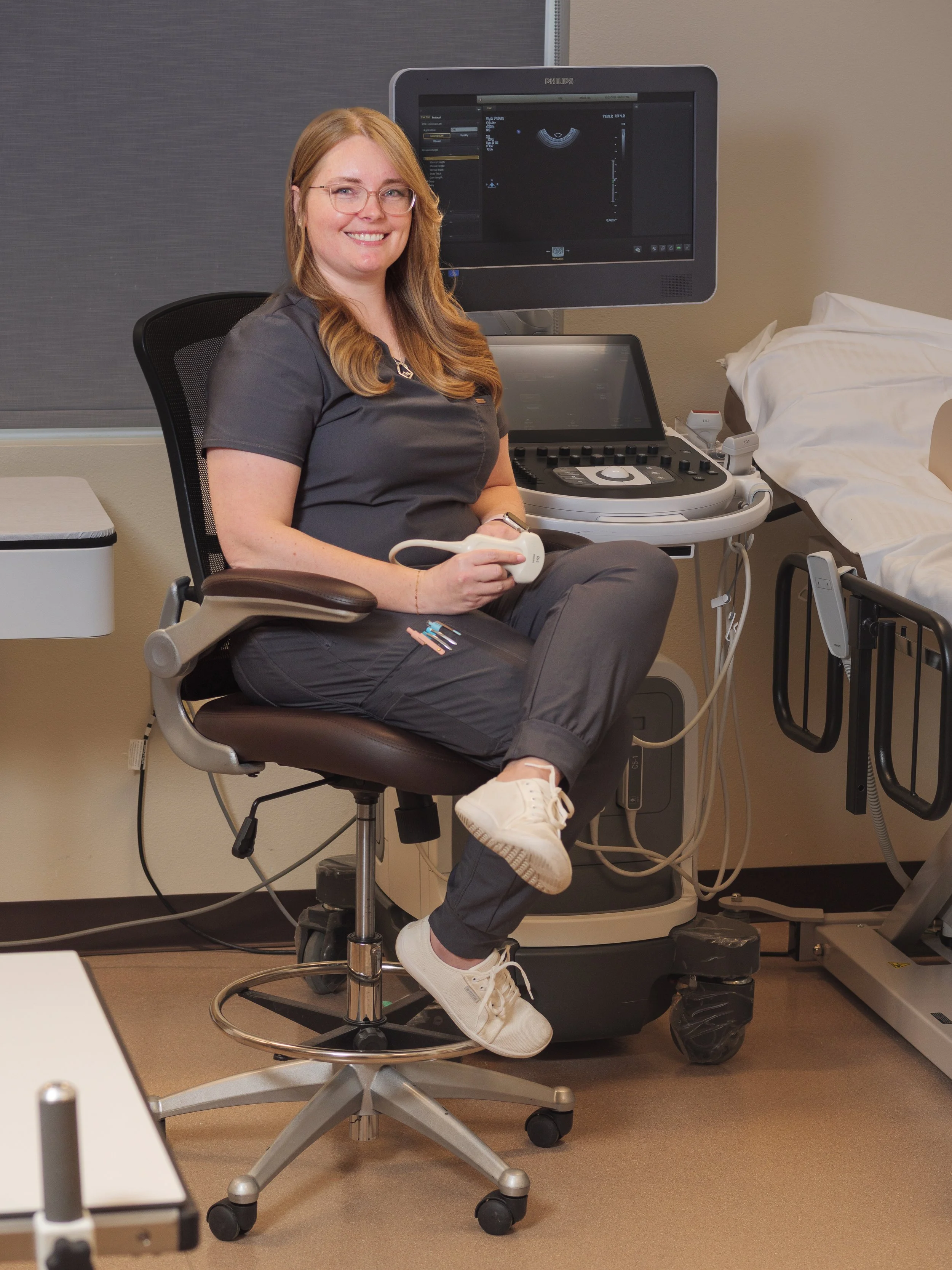 A woman sitting on a medical stool in a healthcare setting, holding an ultrasound probe, with ultrasound equipment and a bed nearby.