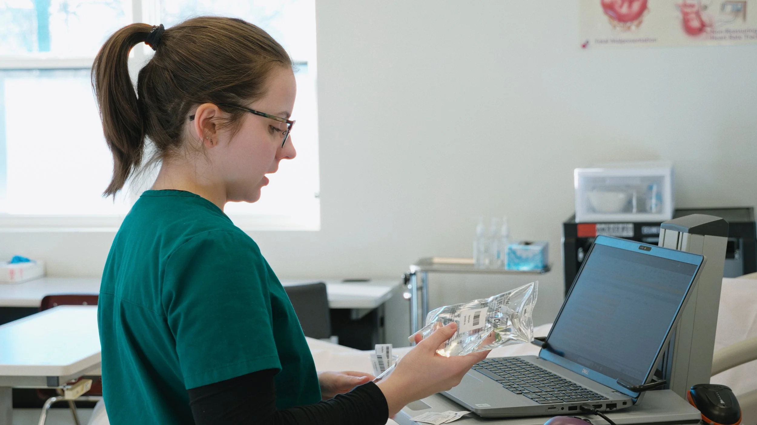 Healthcare worker in scrubs inspecting a sterile medical device in a hospital room with medical supplies and equipment in the background.