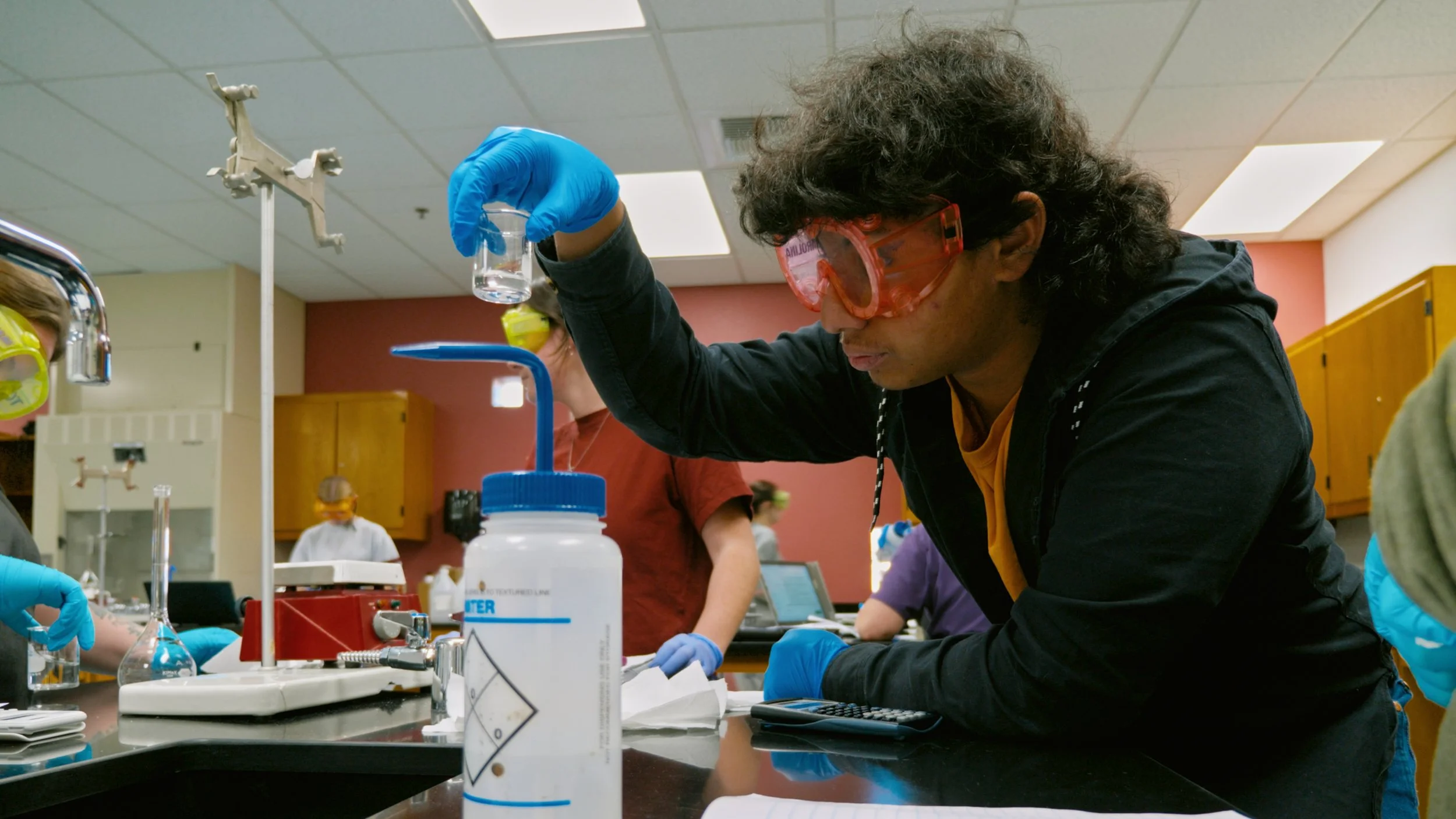 A scientist wearing orange safety goggles and blue gloves conducting an experiment in a laboratory, holding a small beaker above a laboratory table with various lab equipment around.