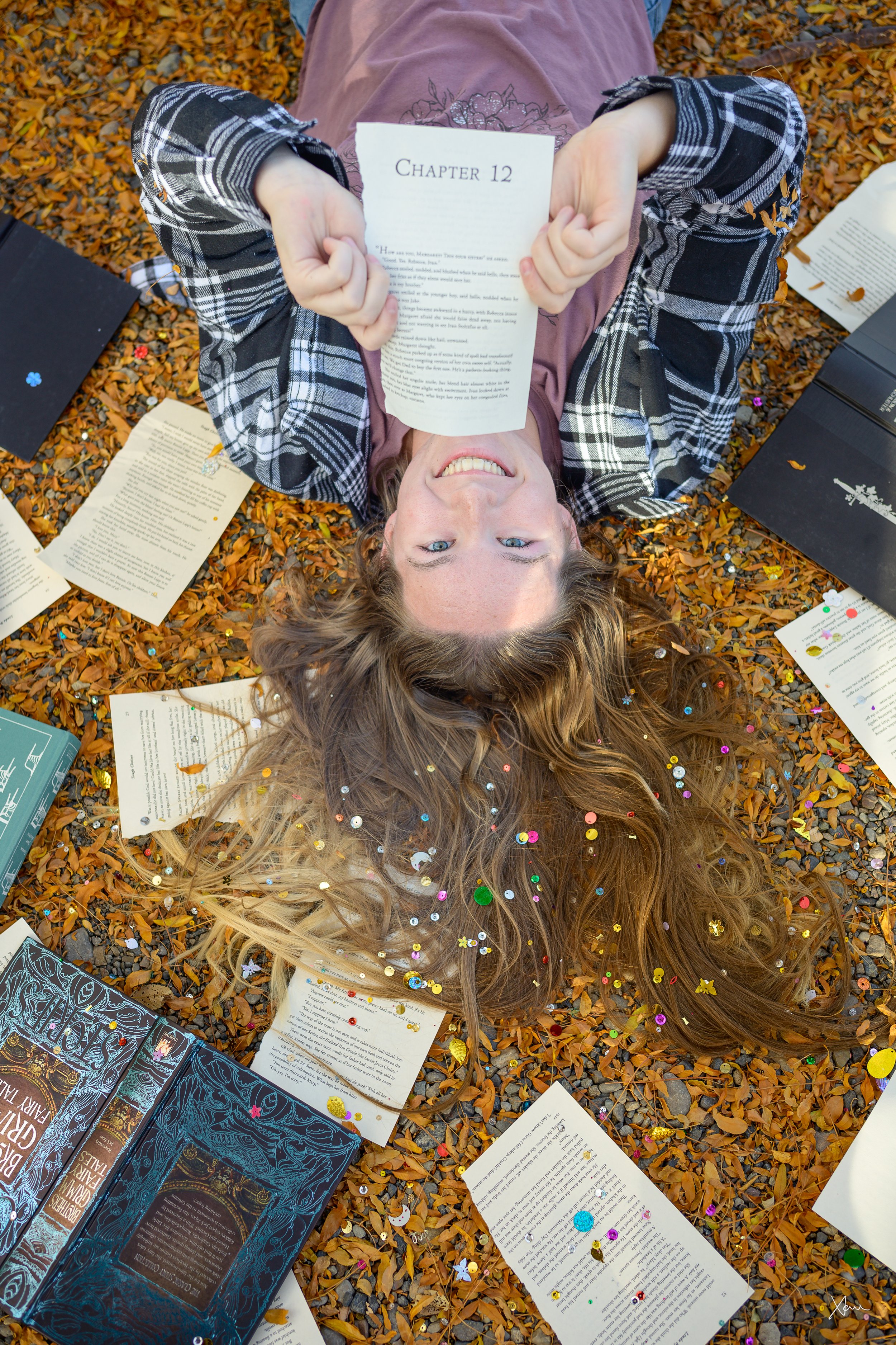 A woman with long wavy hair lies on the ground covered with orange autumn leaves, surrounded by open books, confetti, and a smile while holding a book titled 'Chapter 12' above her face.