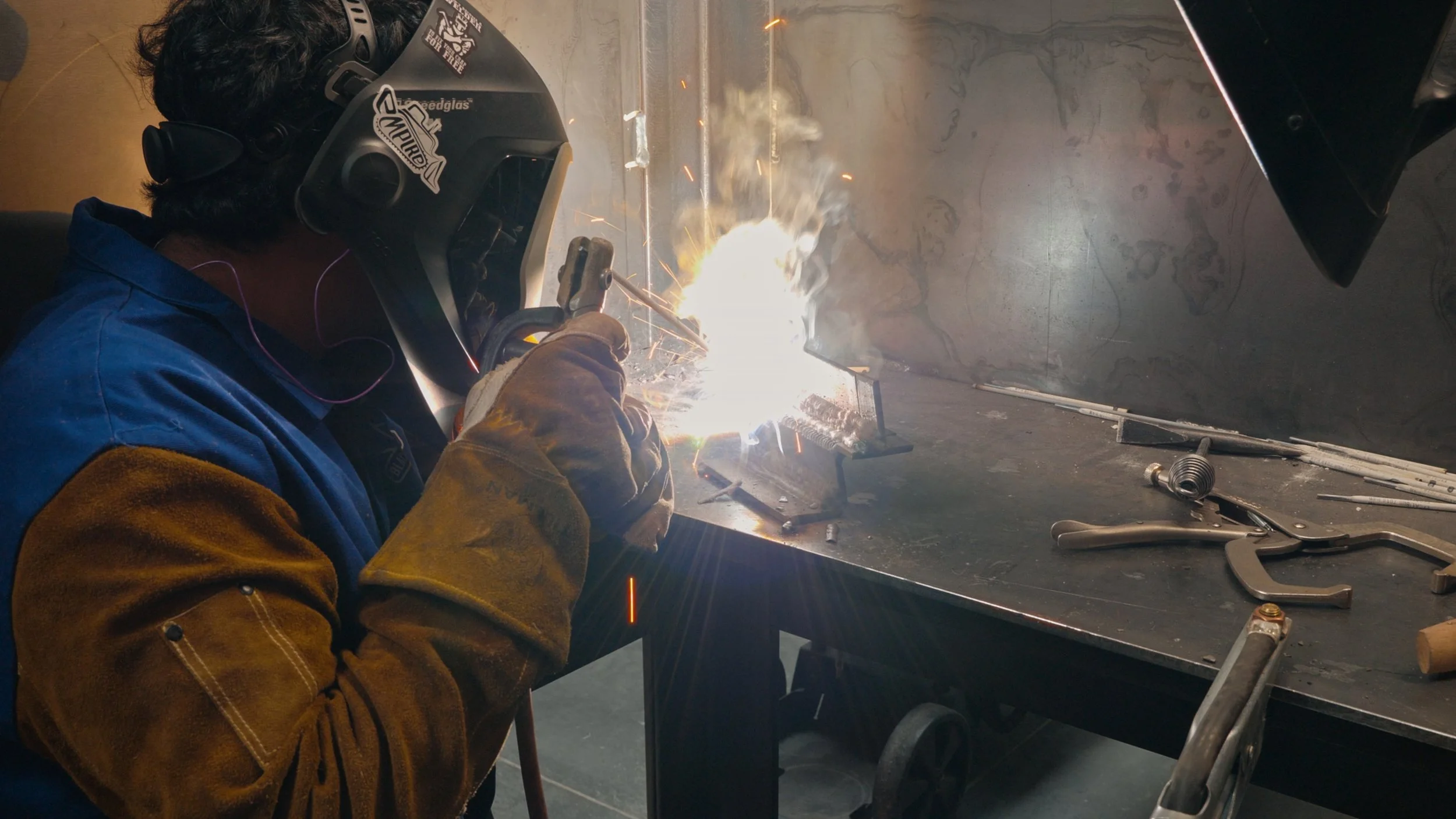 A person welding metal on a workbench, wearing a protective helmet and gloves, with sparks flying from the welding process.