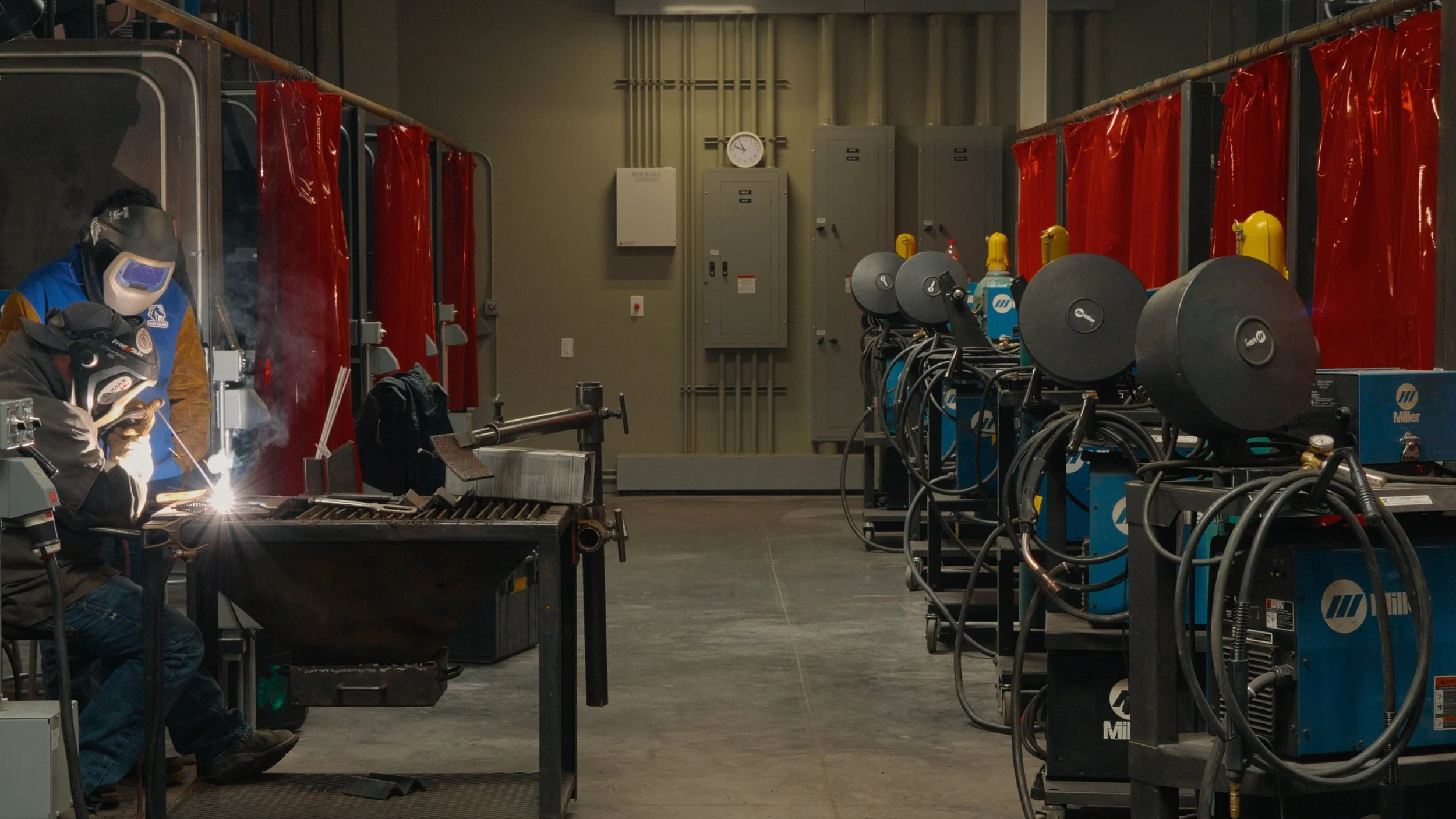 Two individuals welding in a workshop with red safety curtains, welding machines, and electrical panels in the background.