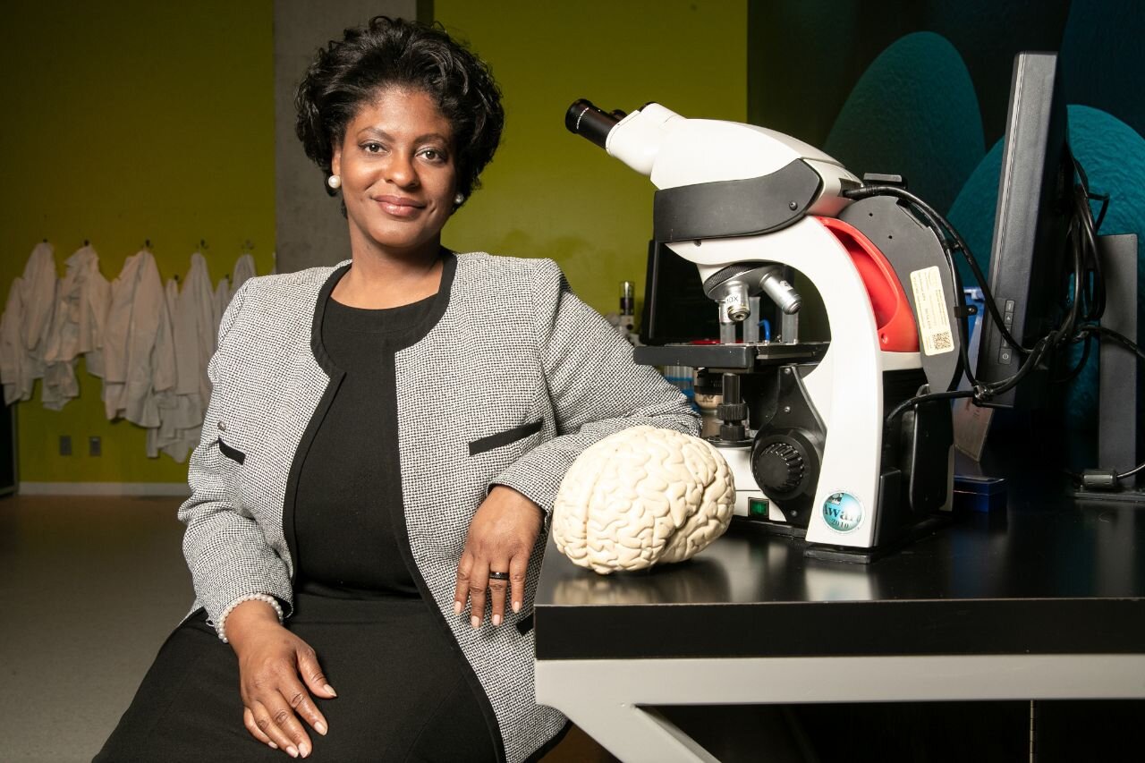 Joyonna Gamble-George in a black dress and gray blazer sitting at a lab bench with a model brain and microscope next to her