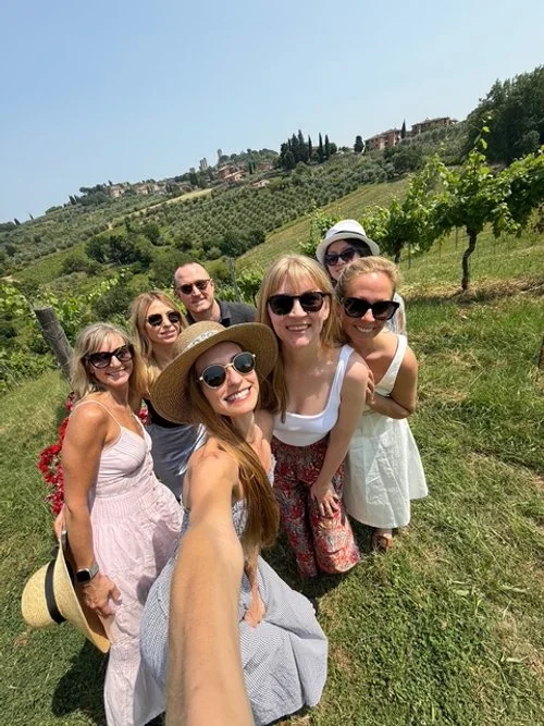 Six women and one man taking a group selfie in a vineyard with rolling hills and houses in the background.