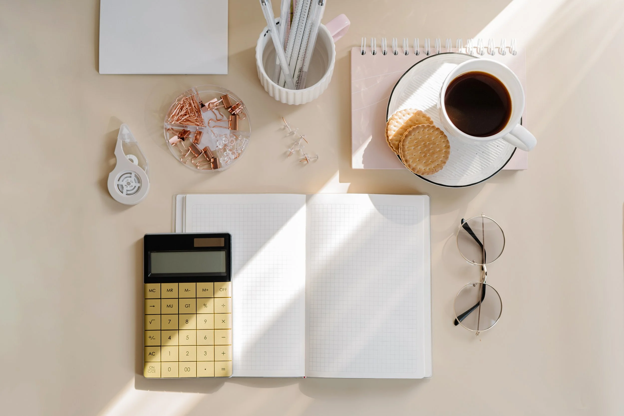 A beige desk with a notebook, calculator, glasses, cup of coffee with crackers, and office supplies.