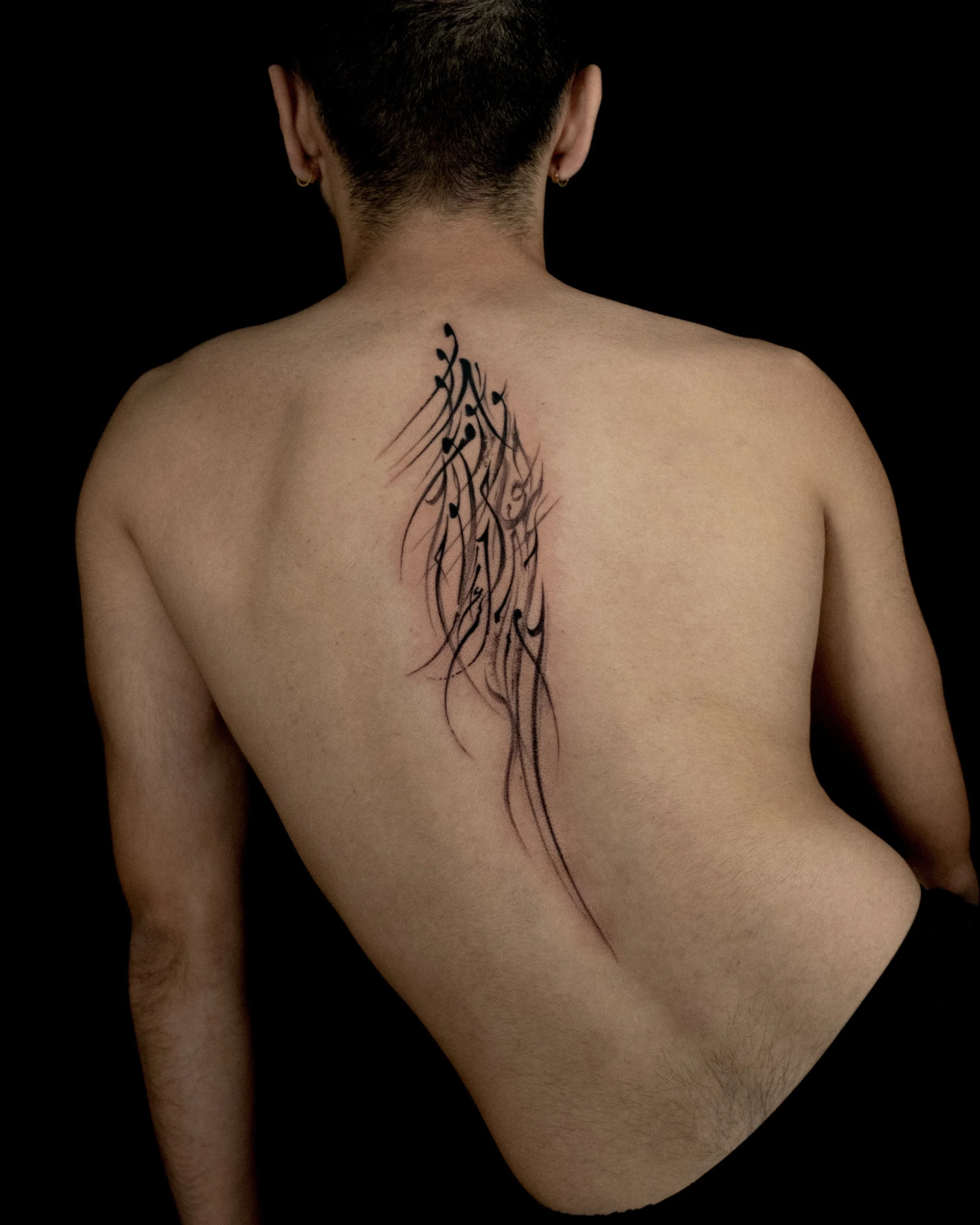 Back view of a person with a vertical black tattoo in a flowing script style on their back.