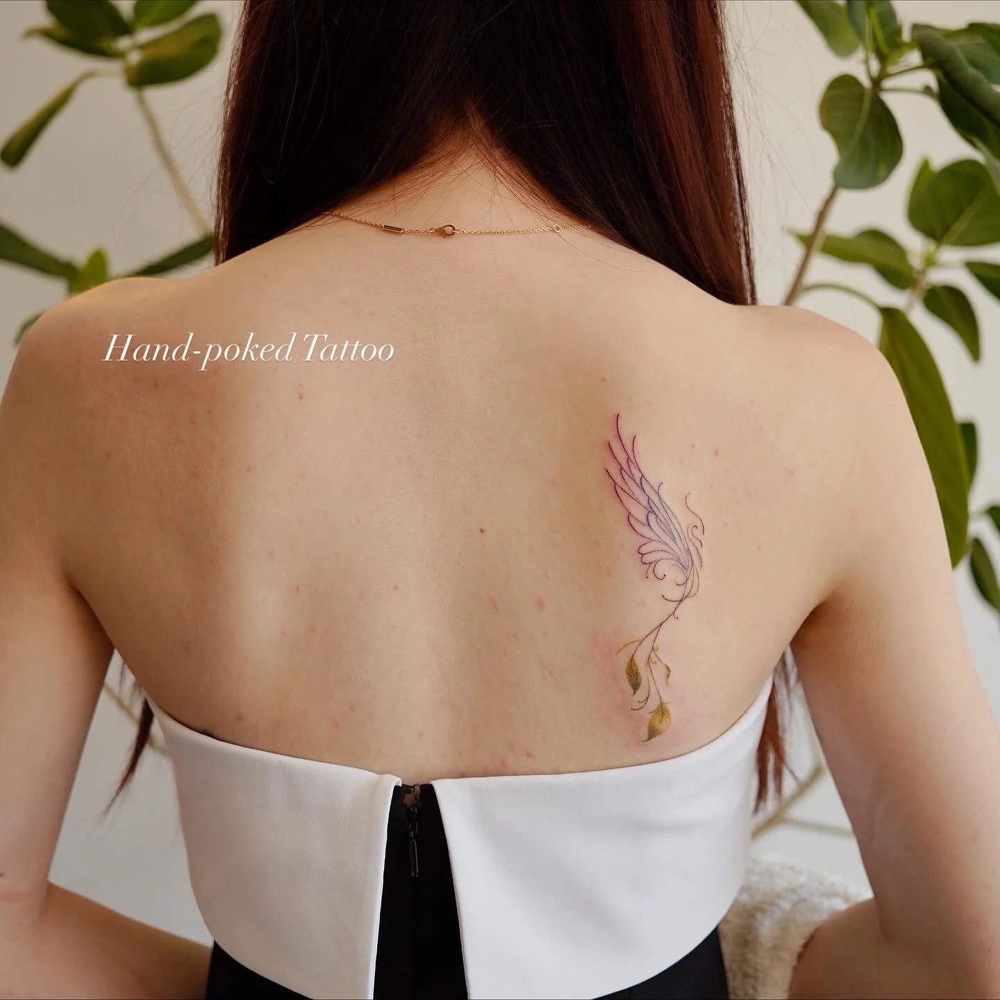 A woman with a tattoo of a winged floral design on her upper back, wearing a strapless white top, with a background of green plants.