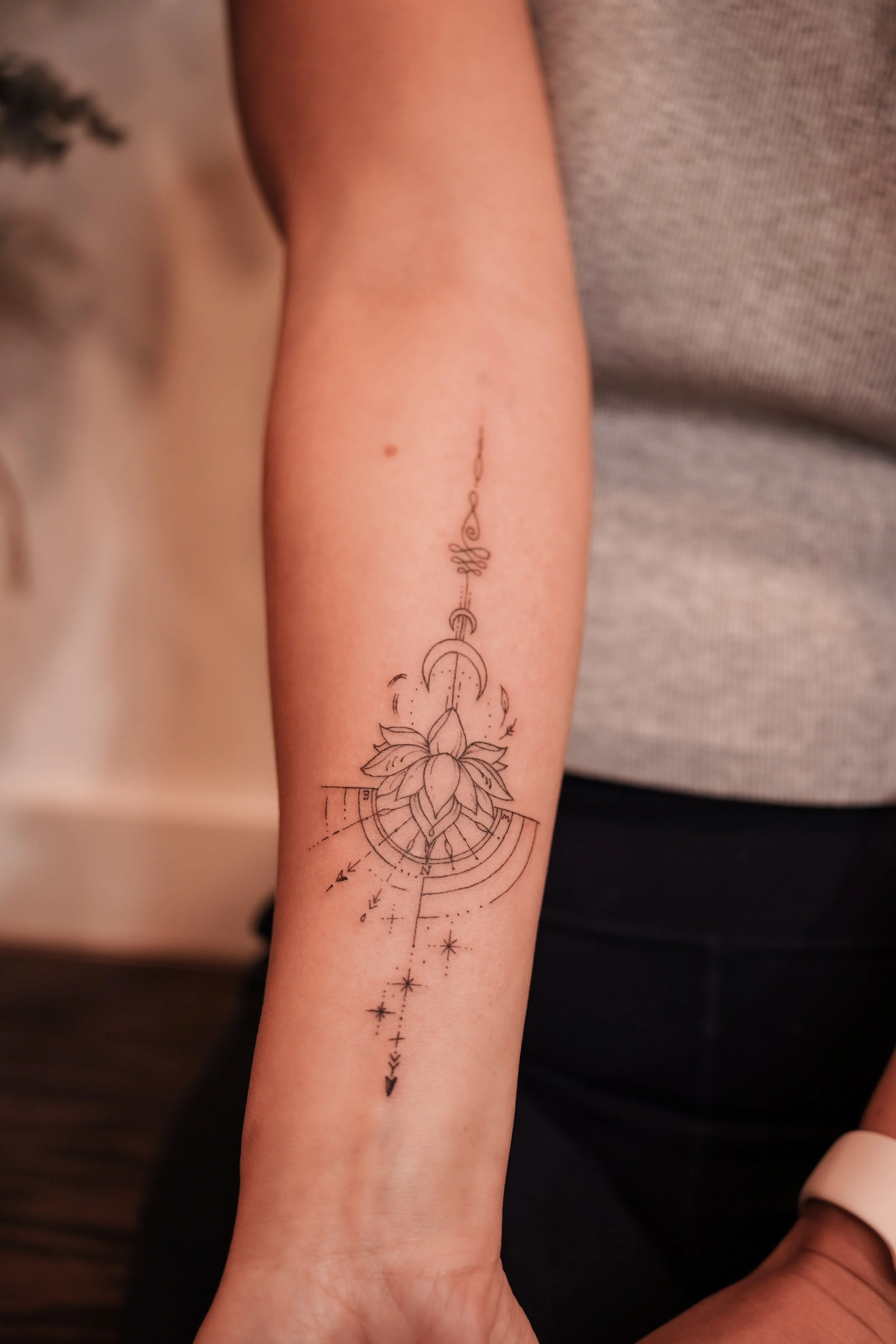 Minimalist line art tattoo on forearm featuring a lotus flower, geometric shapes, crescent moon, and stars.