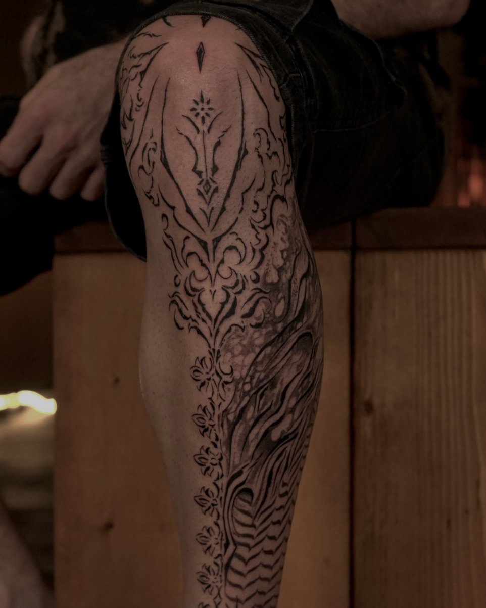 Close-up of a person's leg with an intricate black tribal tattoo covering the thigh and calf, with visible hand resting on a surface in the background.