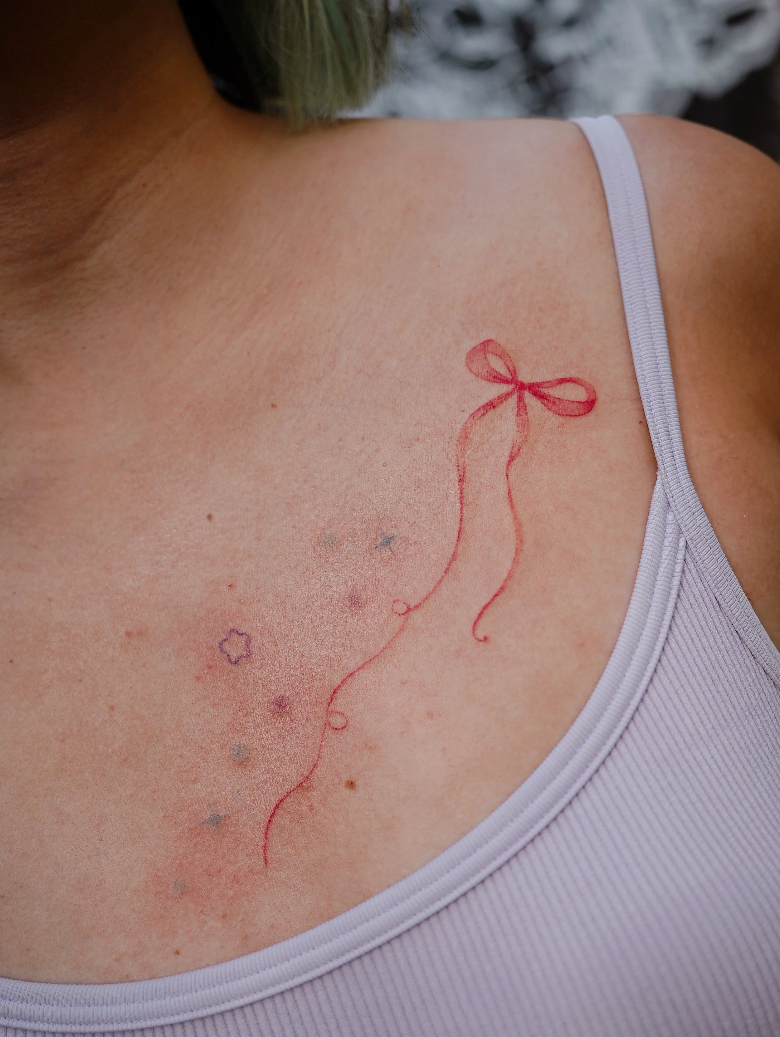 Close-up of a person's shoulder and chest area showing temporary tattoos including a red bow, stars, and circles on light skin and a purple ribbed tank top strap.
