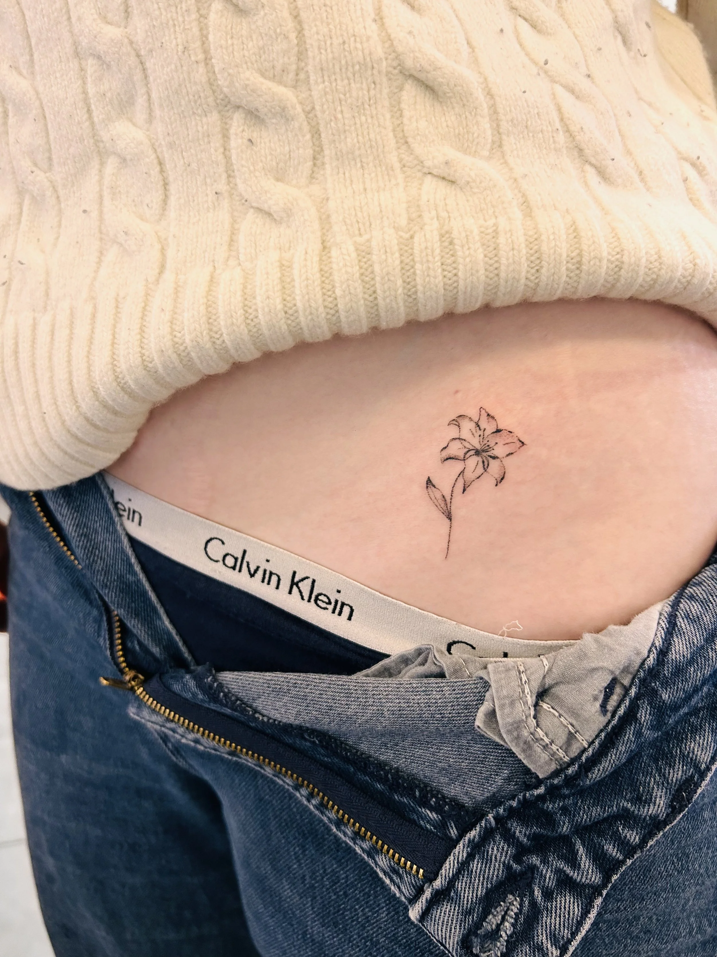 A floral tattoo on the lower abdomen of a person, partially visible above unzipped jeans and a Calvin Klein waistband, with a cream-colored cable-knit sweater.