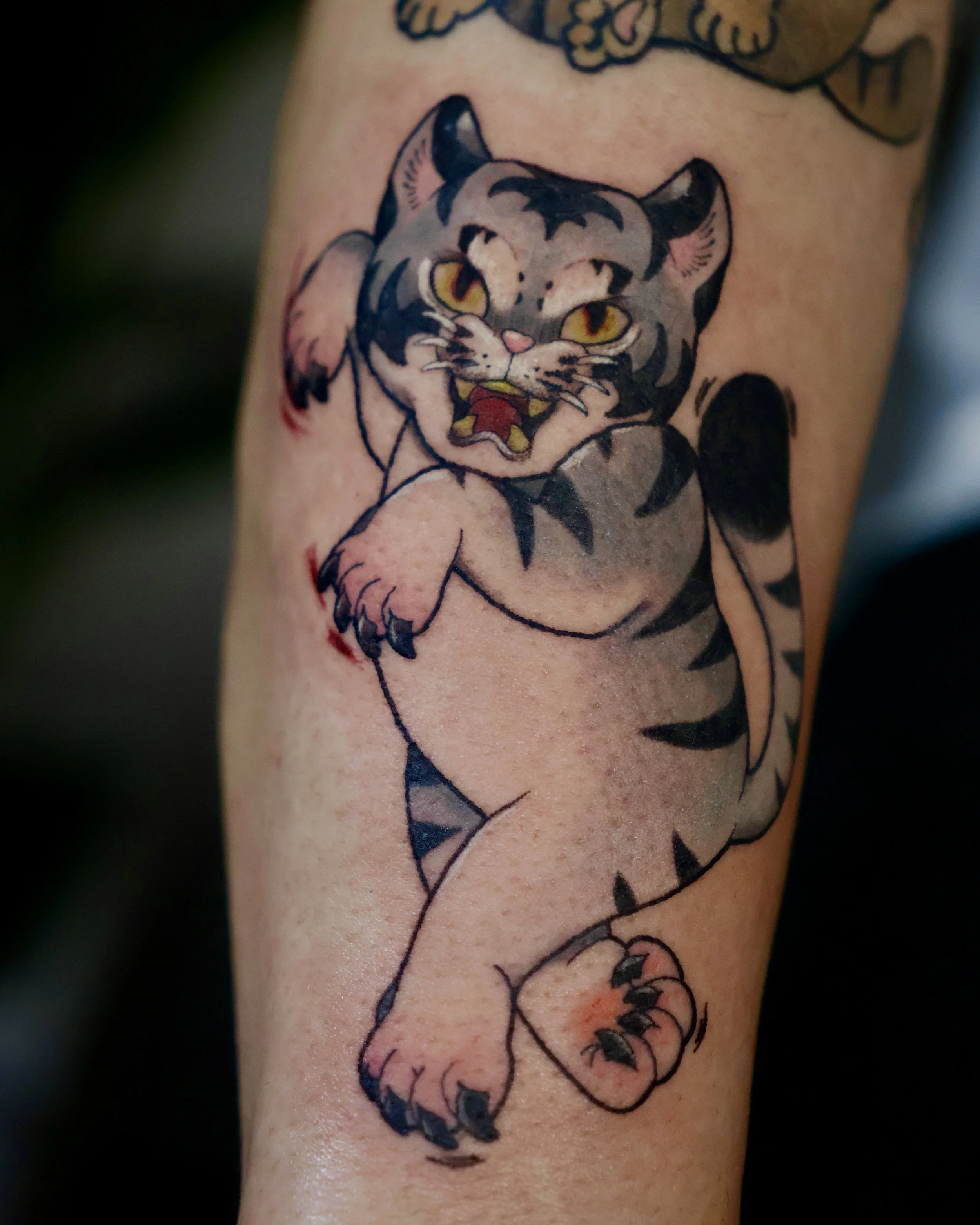Tattoo of a stylized fighting cat with yellow eyes, pink nose, and black stripes, mid-pounce, on person's arm.