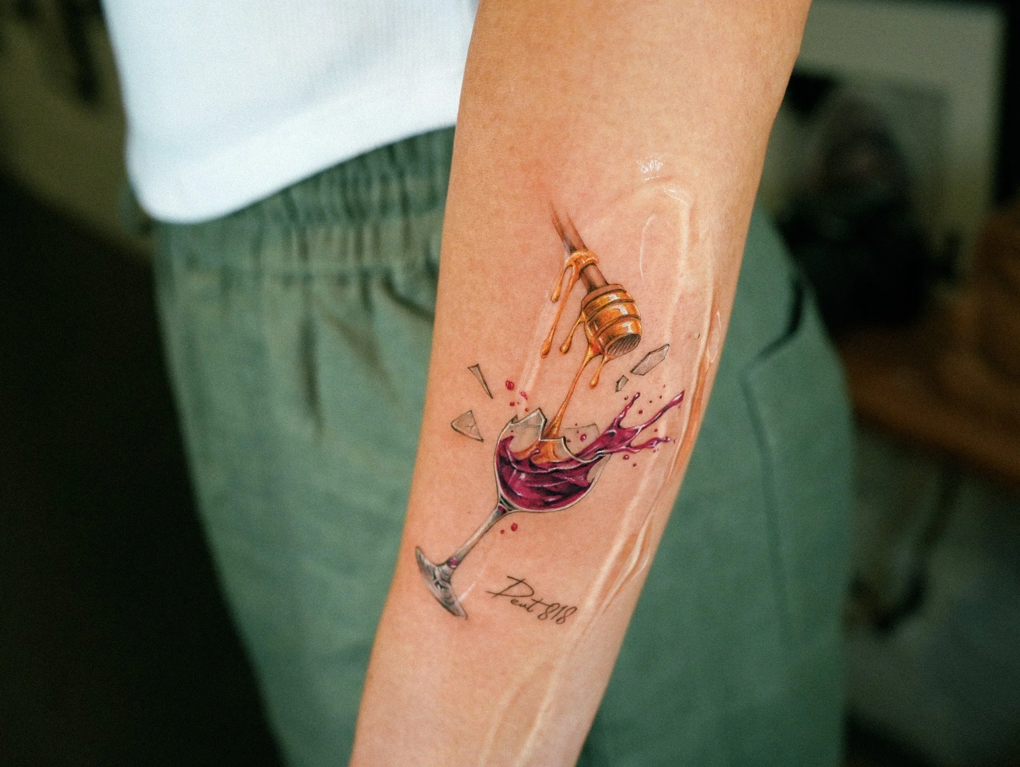 Tattoo of a wine glass with red wine splashing and a honey dipper dripping honey, located on a person's forearm.