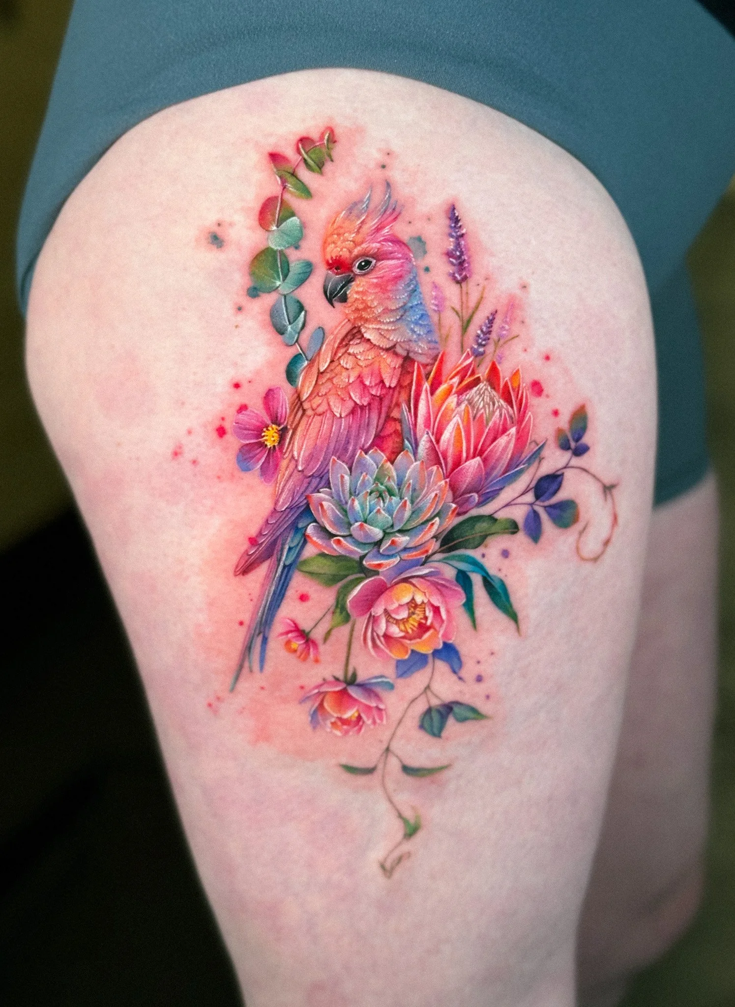 Colorful tattoo of a pink cockatoo perched on large, vibrant pink and purple flowers with green leaves on a person's thigh.