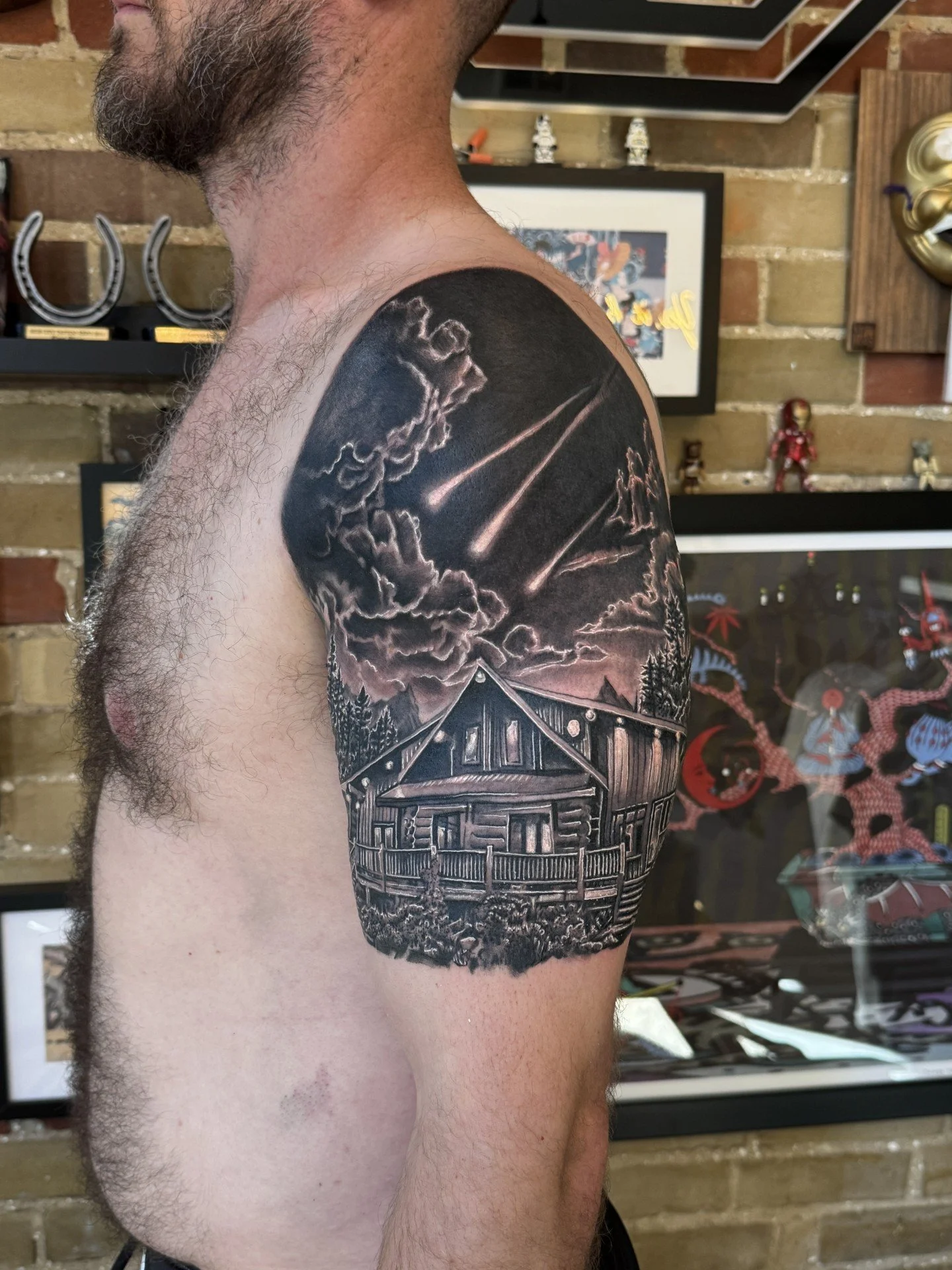 Black and grey realistic tattoo of a log cabin under a night sky with clouds and shooting stars, on a man's upper arm.