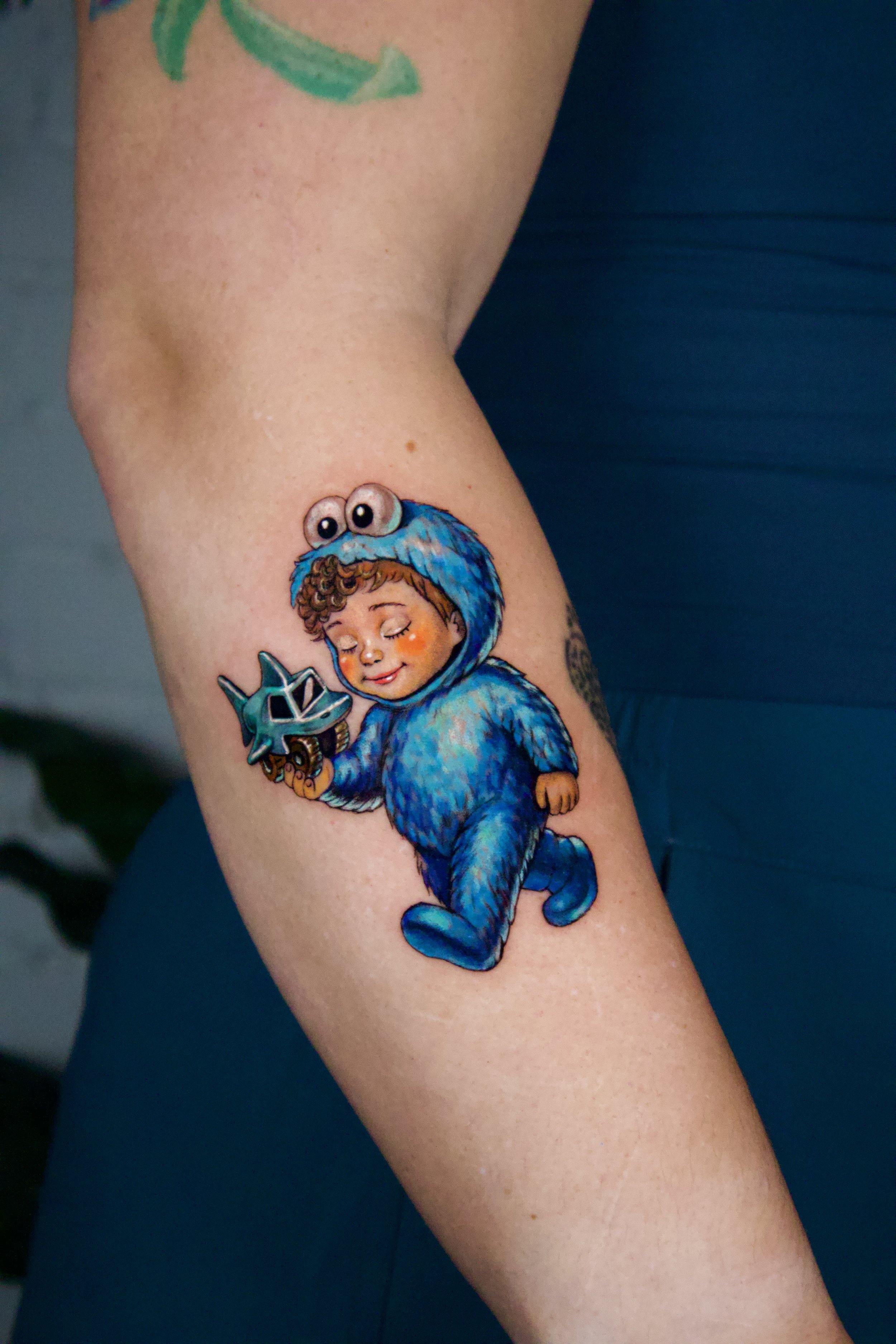 Colorful tattoo of a child in a blue furry costume with googly eyes on the hood, holding a small toy helmet, on a person's arm.