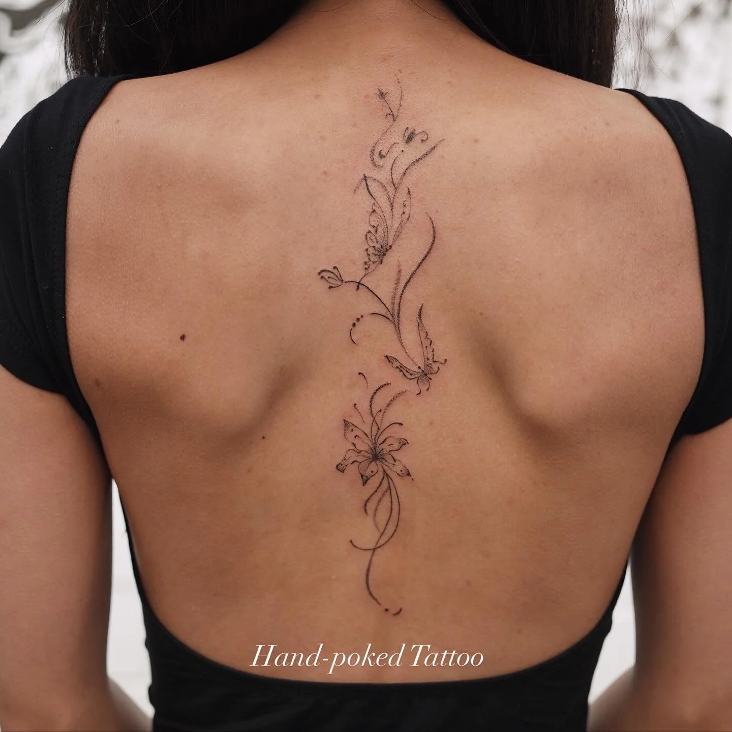 A woman with a black T-shirt showing a delicate vertical floral and butterfly tattoo in black ink on her back. The tattoo includes intricate line work and fine details, with a caption reading 'Hand-poked Tattoo' at the bottom.