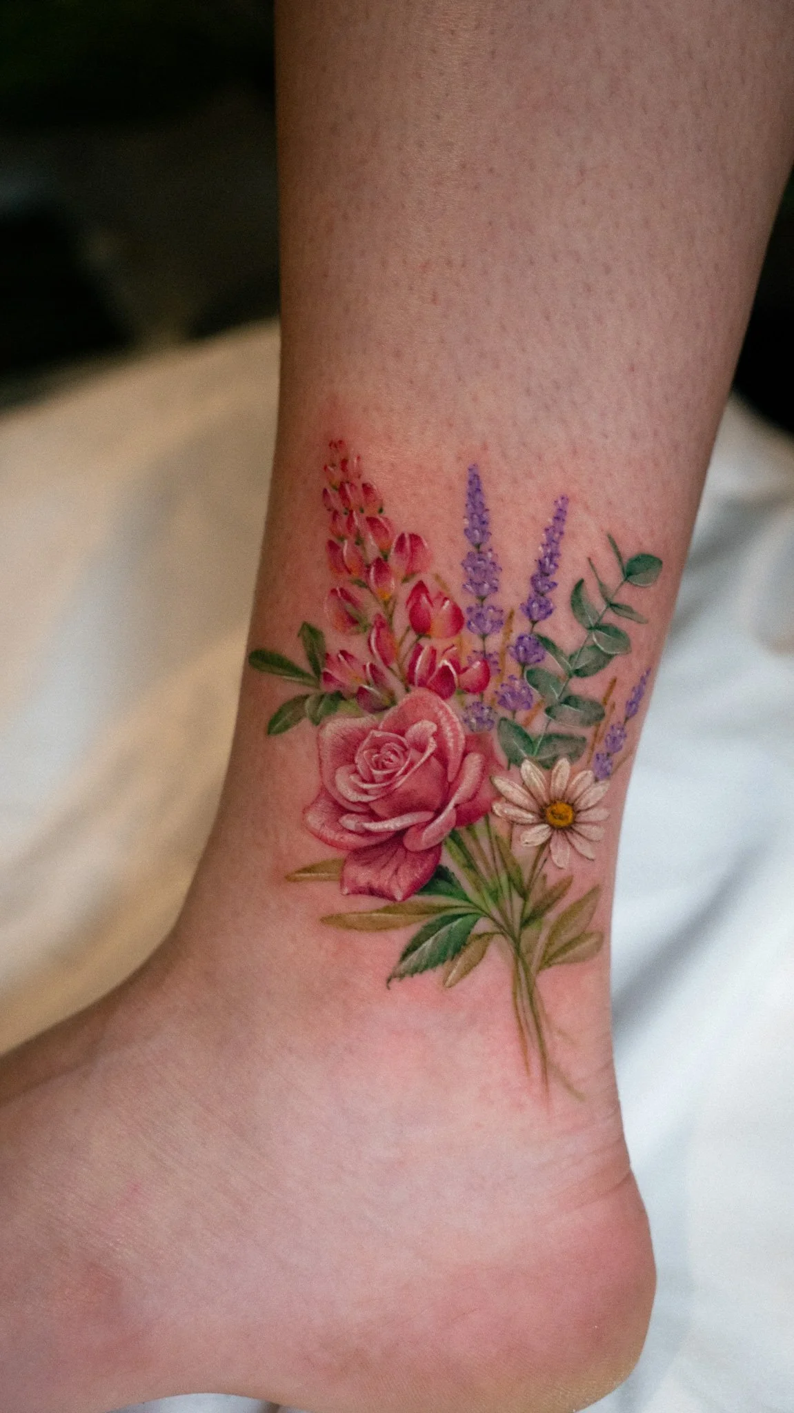 Tattoo of pink roses, purple lavender, daisies, and green leaves on a person's ankle.