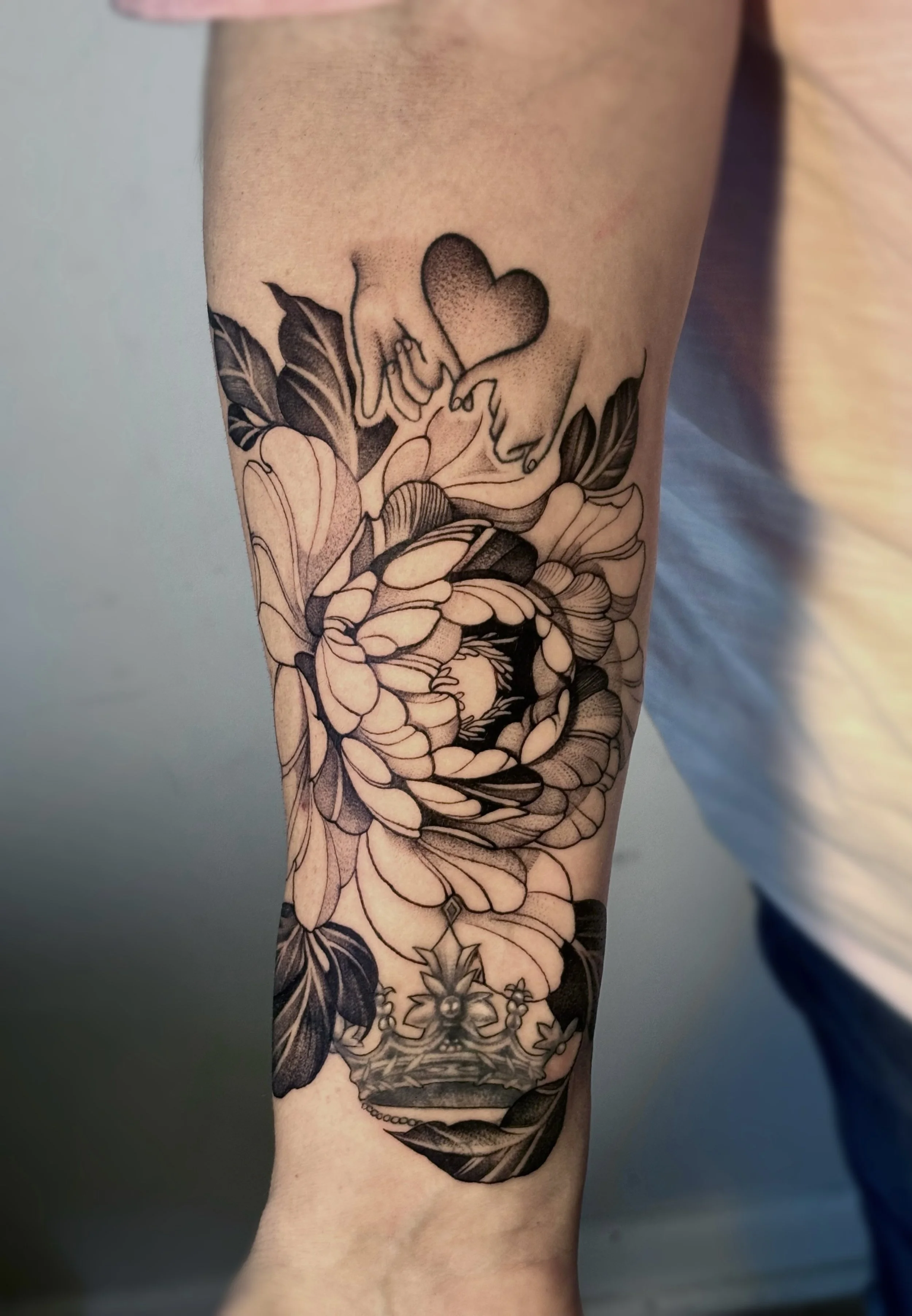 A black and gray tattoo on a person's arm featuring a heart with hands and the word 'Love,' a detailed flower, and a crown at the bottom.