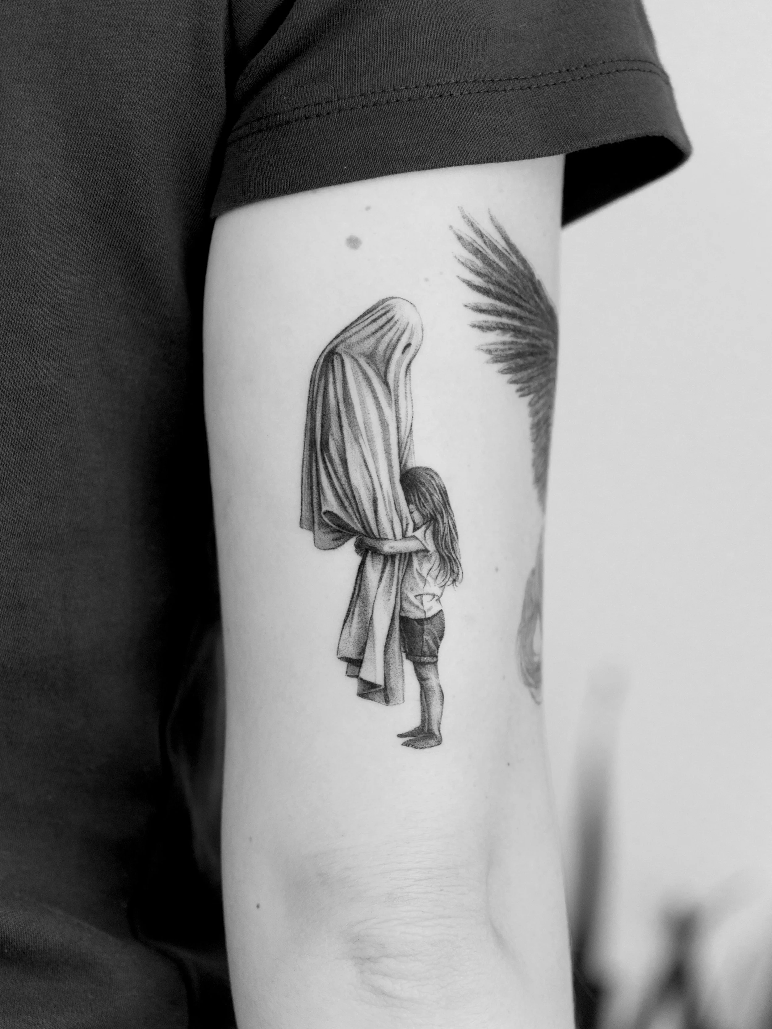 Black and white tattoo of a girl hugging a figure with a veil or hood on her arm, on a person's upper arm.