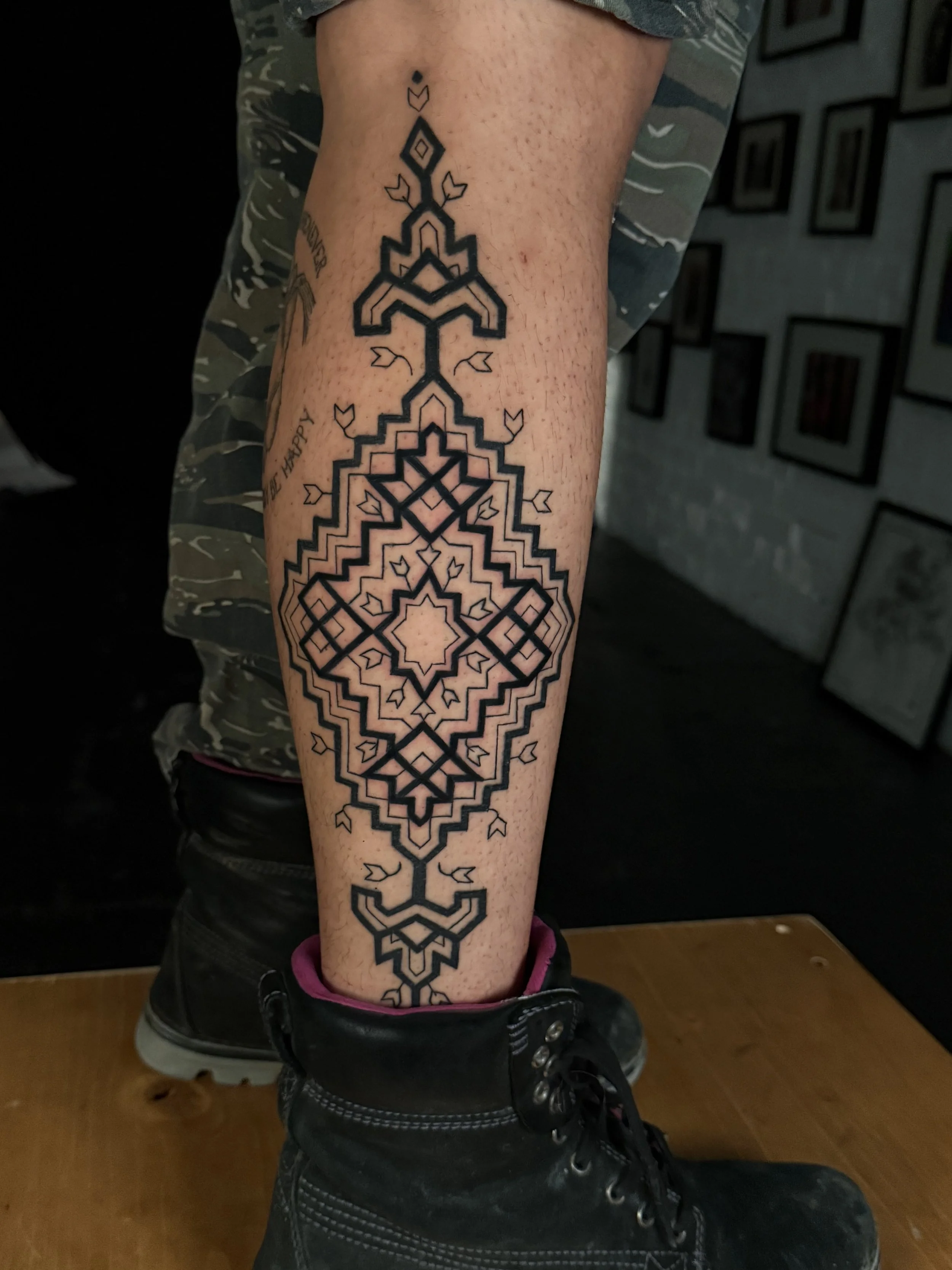 Black geometric tattoo on person's lower leg, featuring symmetrical patterns and small arrow shapes, with the person wearing black combat boots and camo shorts.