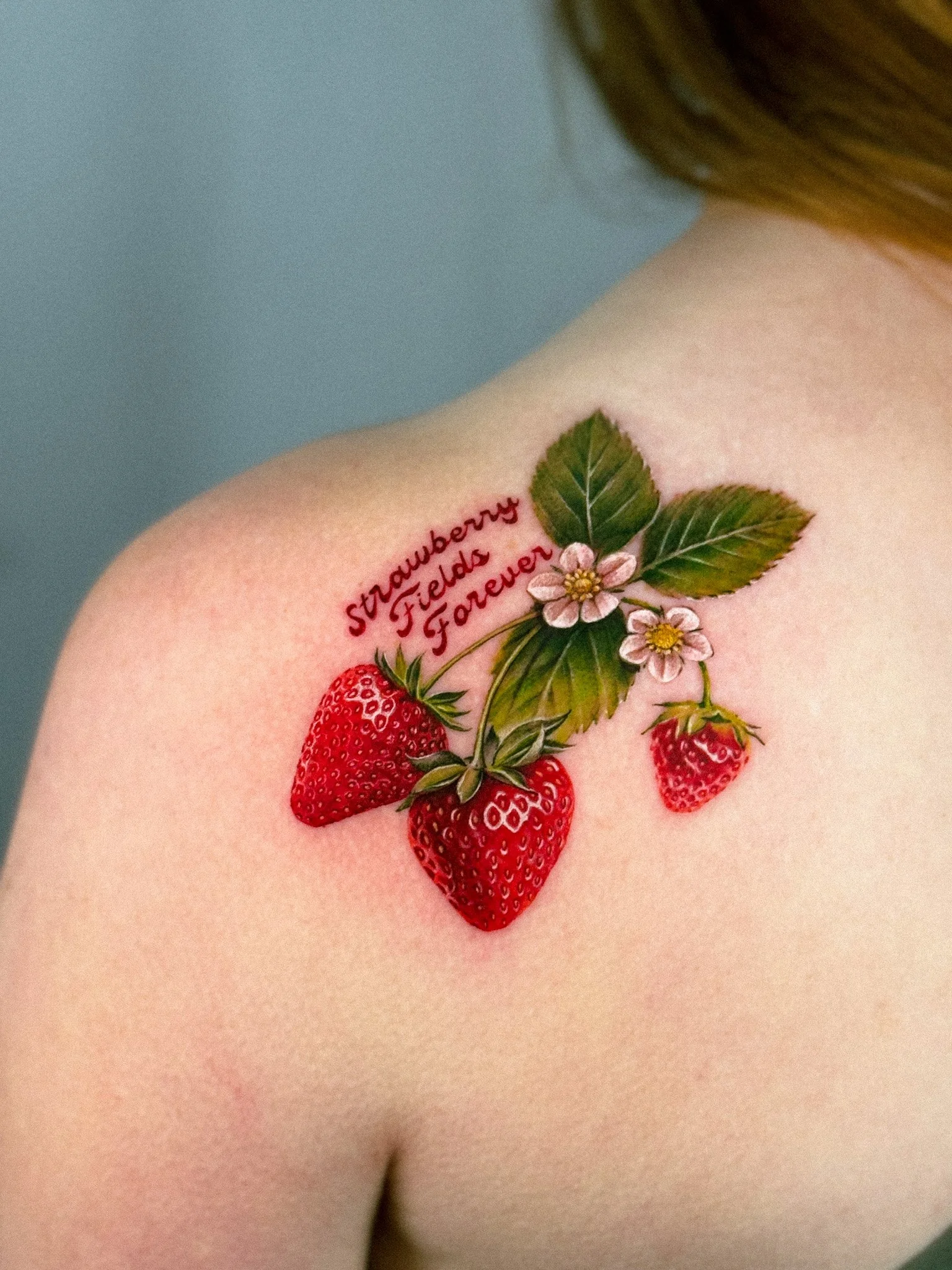 Tattoo of strawberries, leaves, and flowers with the text 'Strawberry Fields Forever' on a person's shoulder.