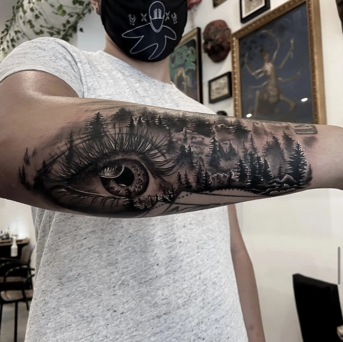 A person with a detailed black and gray tattoo of an eye and a forest scene on their forearm, wearing a gray t-shirt and a black face mask with a design, standing in a room decorated with framed art and masks on the wall.