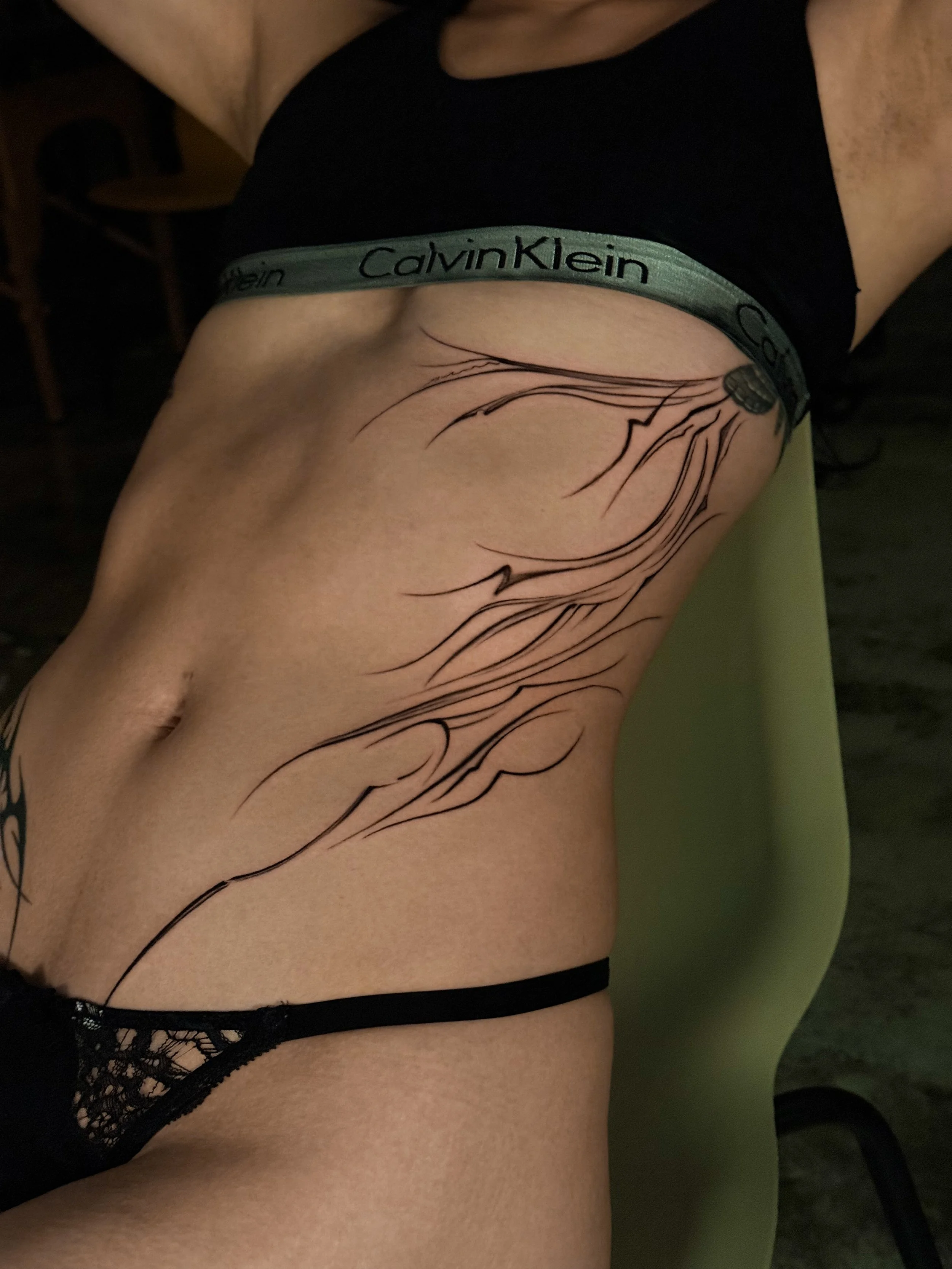 A person lying on a chair showing their side with a tattoo of abstract, flowing lines on their ribcage and abdomen, wearing Calvin Klein underwear.