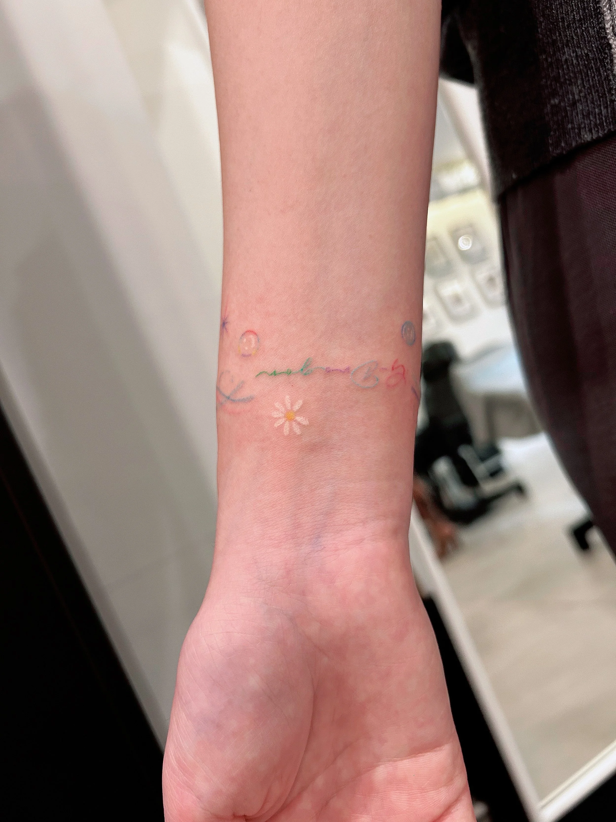 A close-up of a person's inner forearm with a colorful, delicate tattoo featuring the words 'make me feel' in cursive, a small painted daisy, a red heart, and a smiling face.