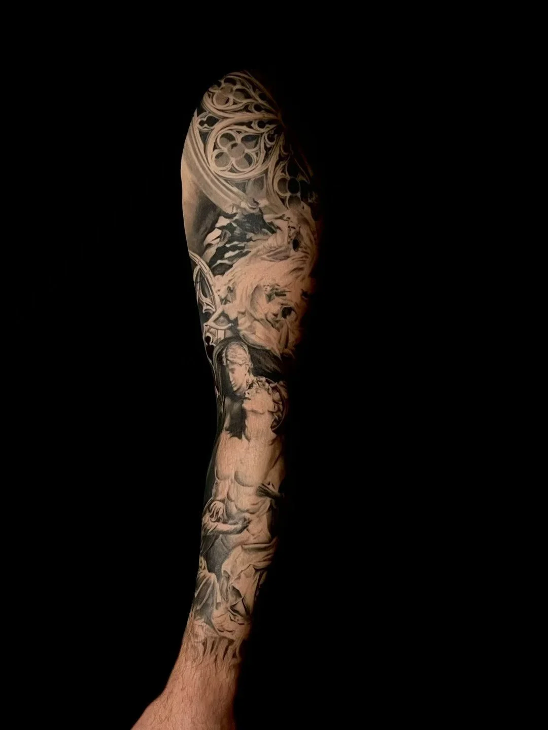 Close-up of a person's arm with detailed black and grey tattoo sleeve depicting various artistic and mythological scenes against a black background.