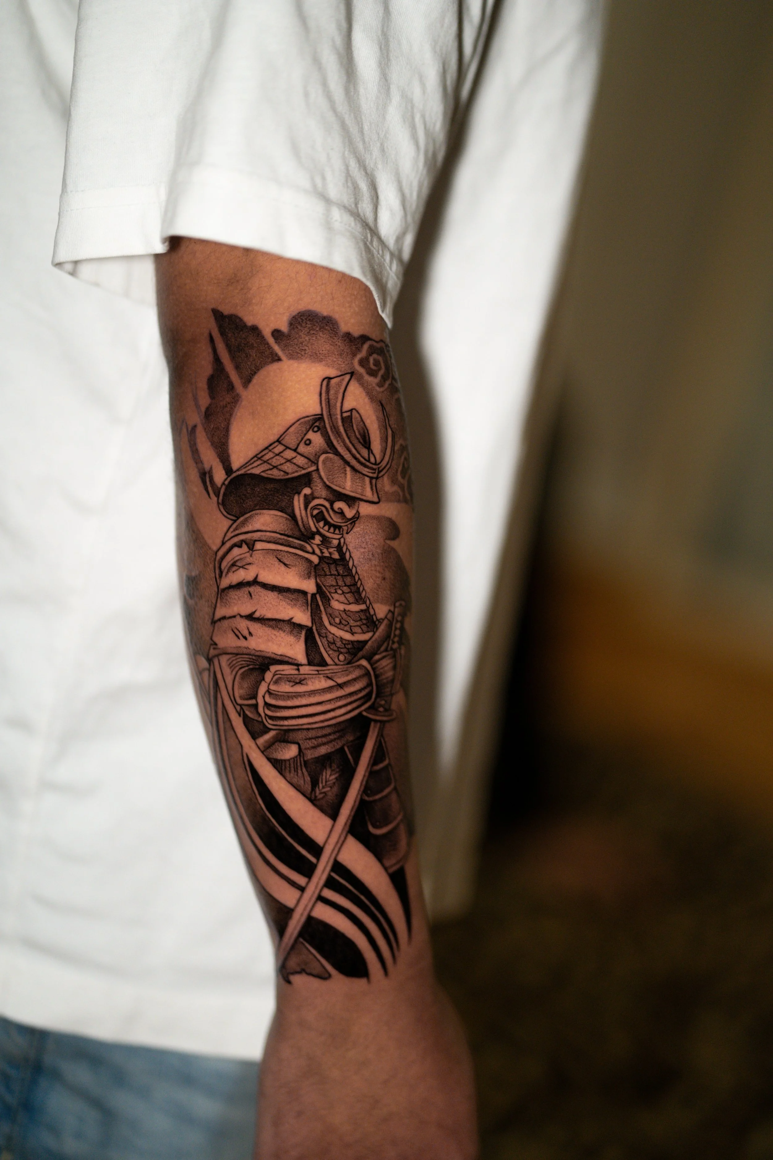 Black and grey tattoo of a samurai warrior holding a sword, with clouds and mountains in the background on a person's arm.