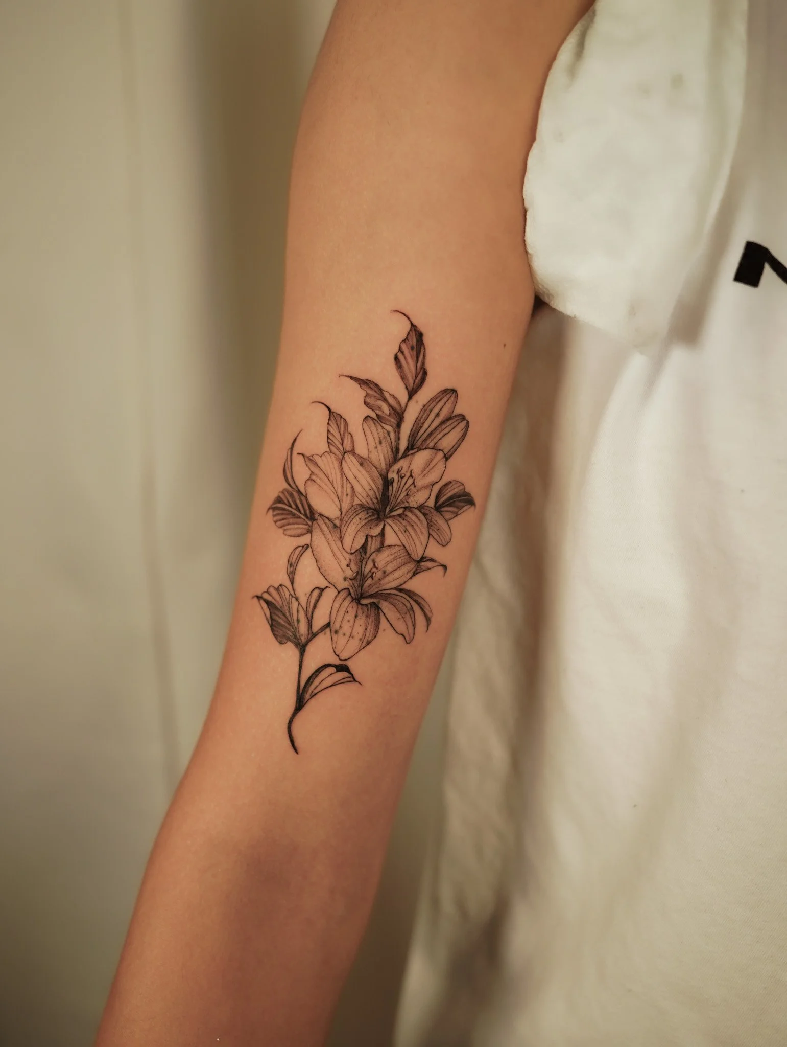 Black outline tattoo of lilies on a person's inner forearm, next to a white sleeveless shirt.